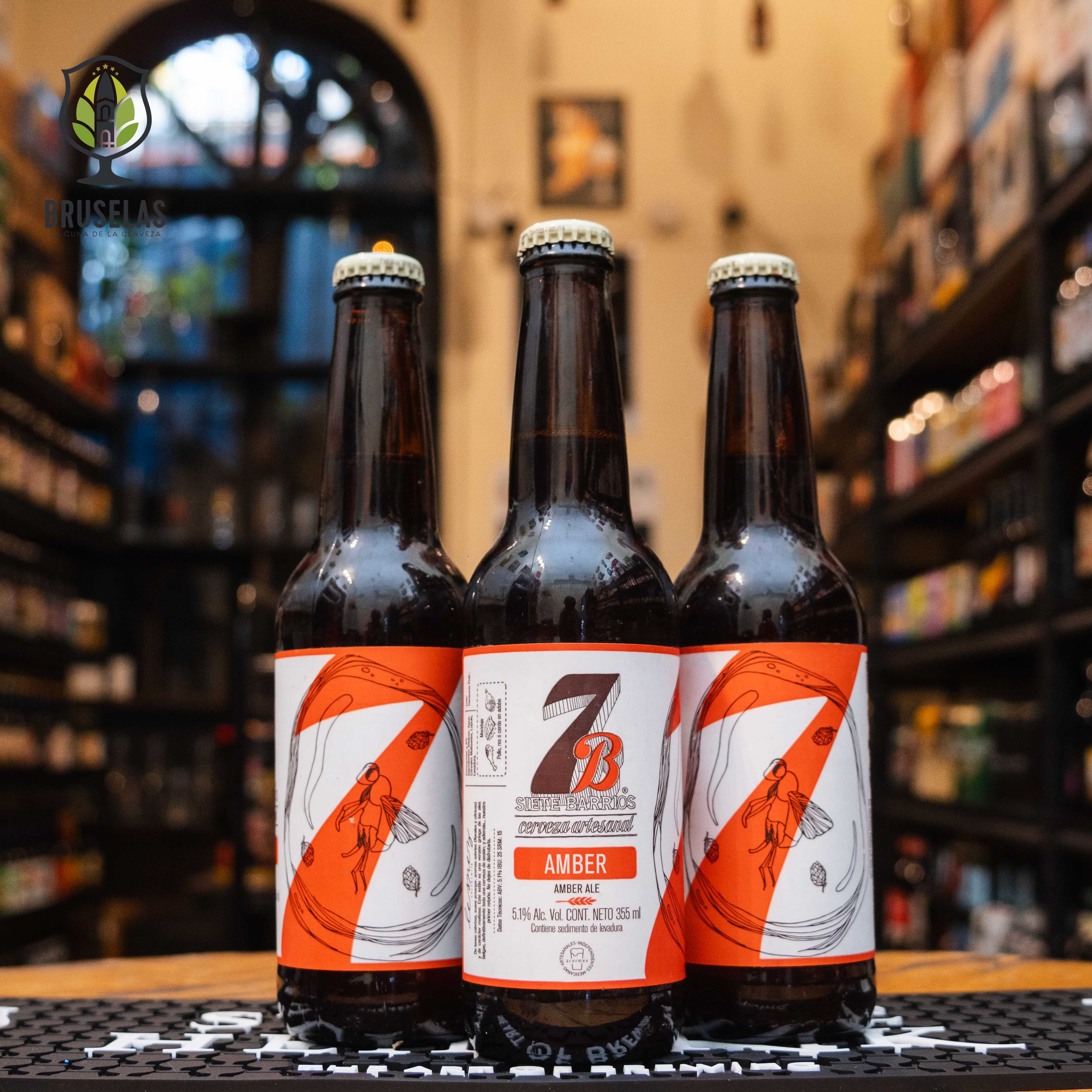 Botella de 7 Barrios Amber Ale, una cerveza artesanal de Cervecería 7 Barrios en San Luis Potosí. La etiqueta es blanca con detalles en rojo y marrón, y el nombre en letras marrones. ABV de 6.1%, con un sabor profundo a malta y aromas florales y cítricos. Ideal para maridar con pollo, res o cerdo con adobos de pungencia media. El diseño refleja la dedicación artesanal de esta cervecería.