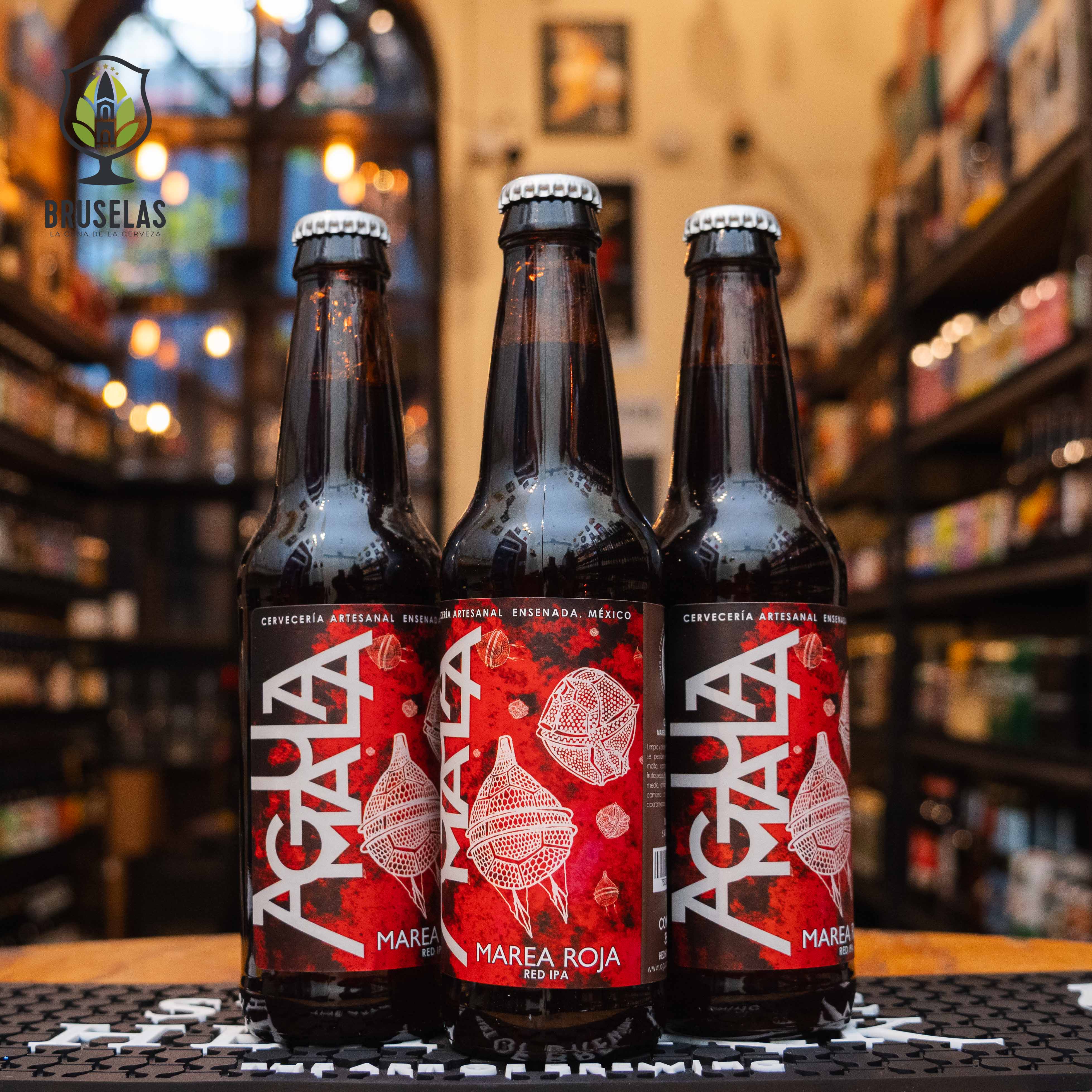 Botella de Agua Mala Marea Roja, una Red IPA elaborada por Cervecería Agua Mala en Ensenada, Baja California. La etiqueta roja muestra un diseño con medusas blancas. ABV de 5.4%, aroma a caramelo y malta tostada con un toque de lúpulo. Ideal para acompañar carnes a la parrilla y quesos maduros. El diseño refleja la naturaleza intensa y equilibrada de esta cerveza.