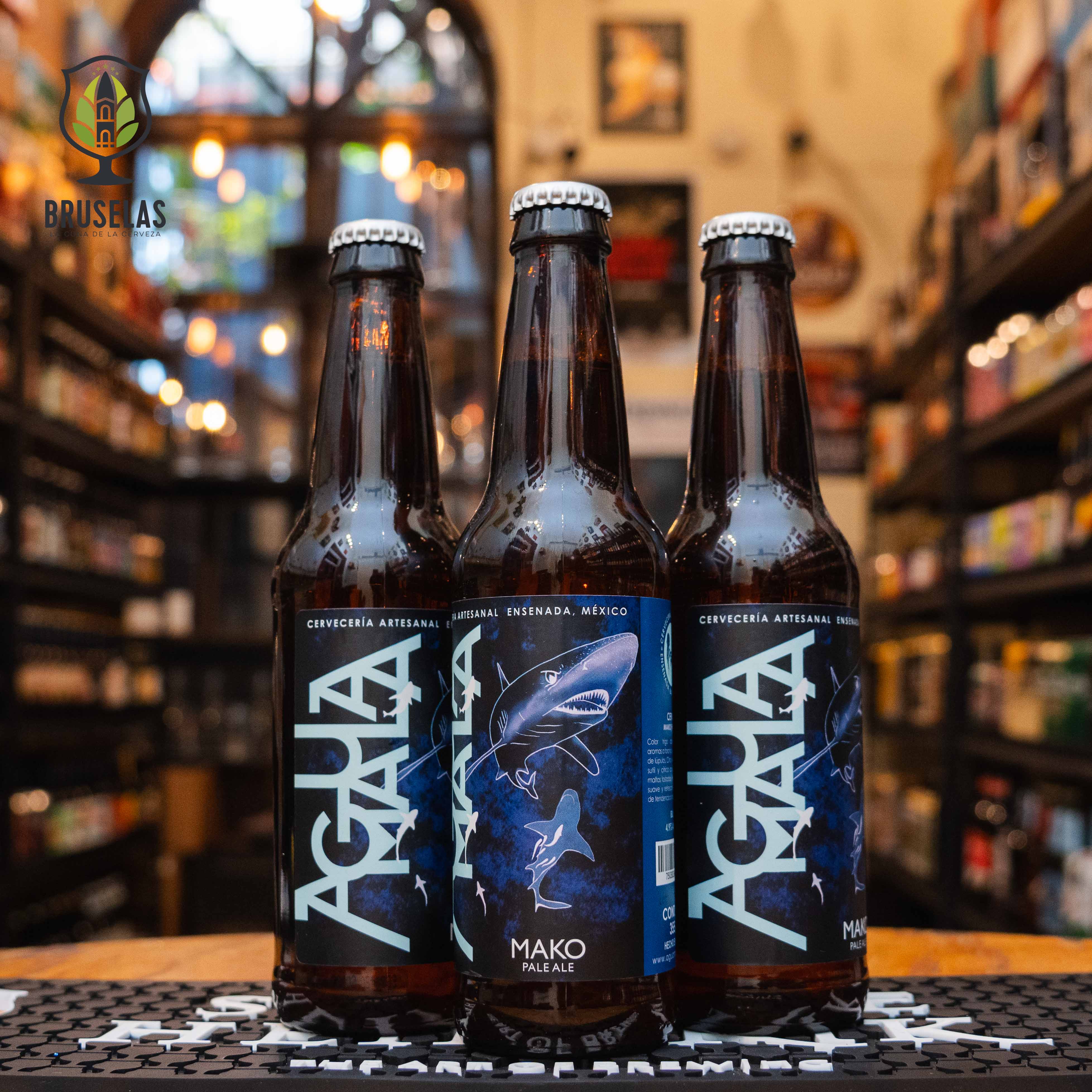 Botella de Agua Mala Mako, una American Pale Ale (APA) de Cervecería Agua Mala en Ensenada, Baja California. La etiqueta presenta un tiburón sobre un fondo azul oscuro. ABV de 4.9% y aroma cítrico y tropical con notas de pino. Ideal para acompañar mariscos y pescados a la parrilla. El diseño refleja el carácter refrescante y lupulado de esta cerveza.