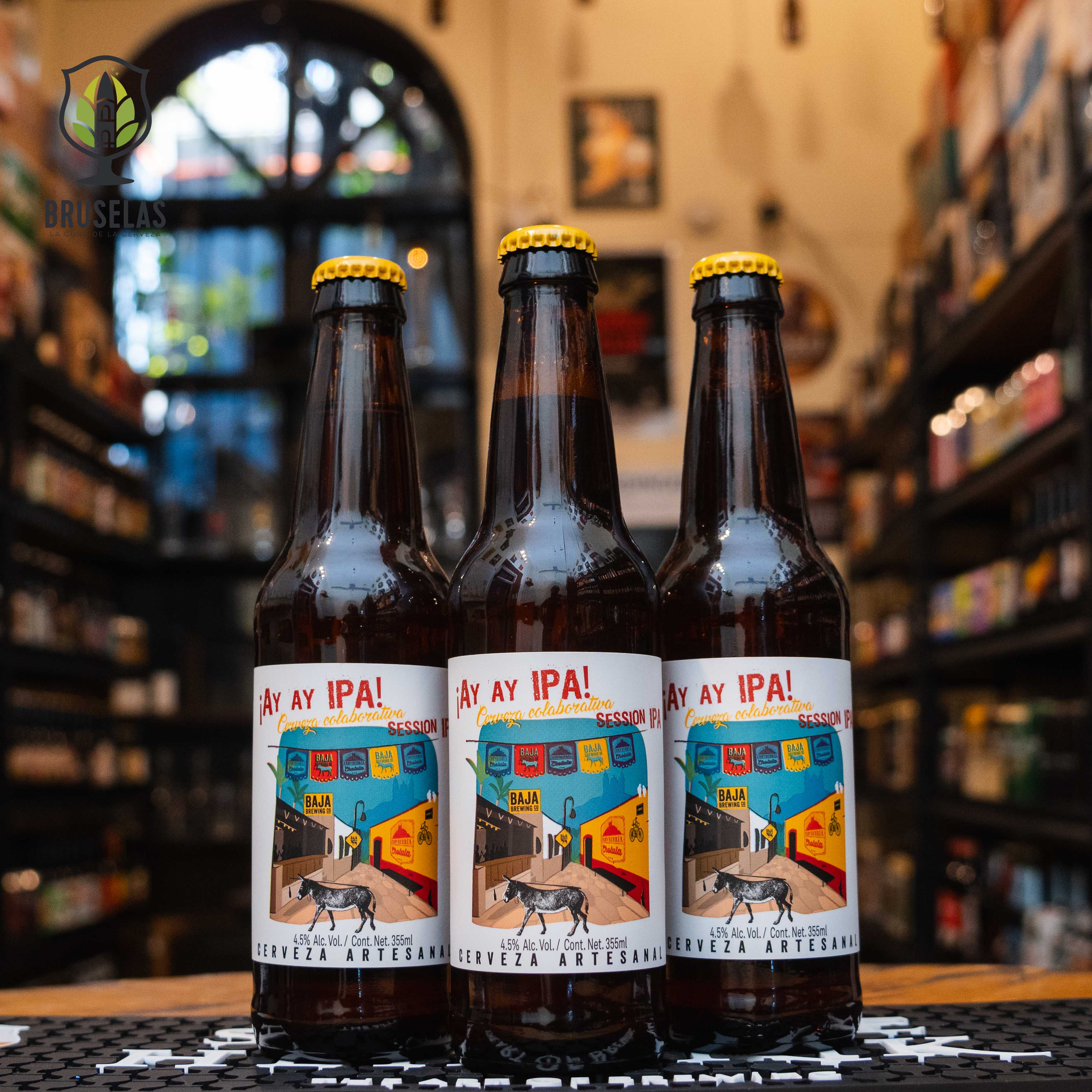 Botella de Baja Ay Ay IPA, una Session IPA colaborativa entre Baja Brewing Co. y Cervecería Cholula. La etiqueta colorida presenta un paisaje urbano con edificios y un perro. ABV de 4.5%, con intensos aromas a lúpulo, cítricos y resina de pino. Ideal para maridar con ensaladas frescas y pizzas.