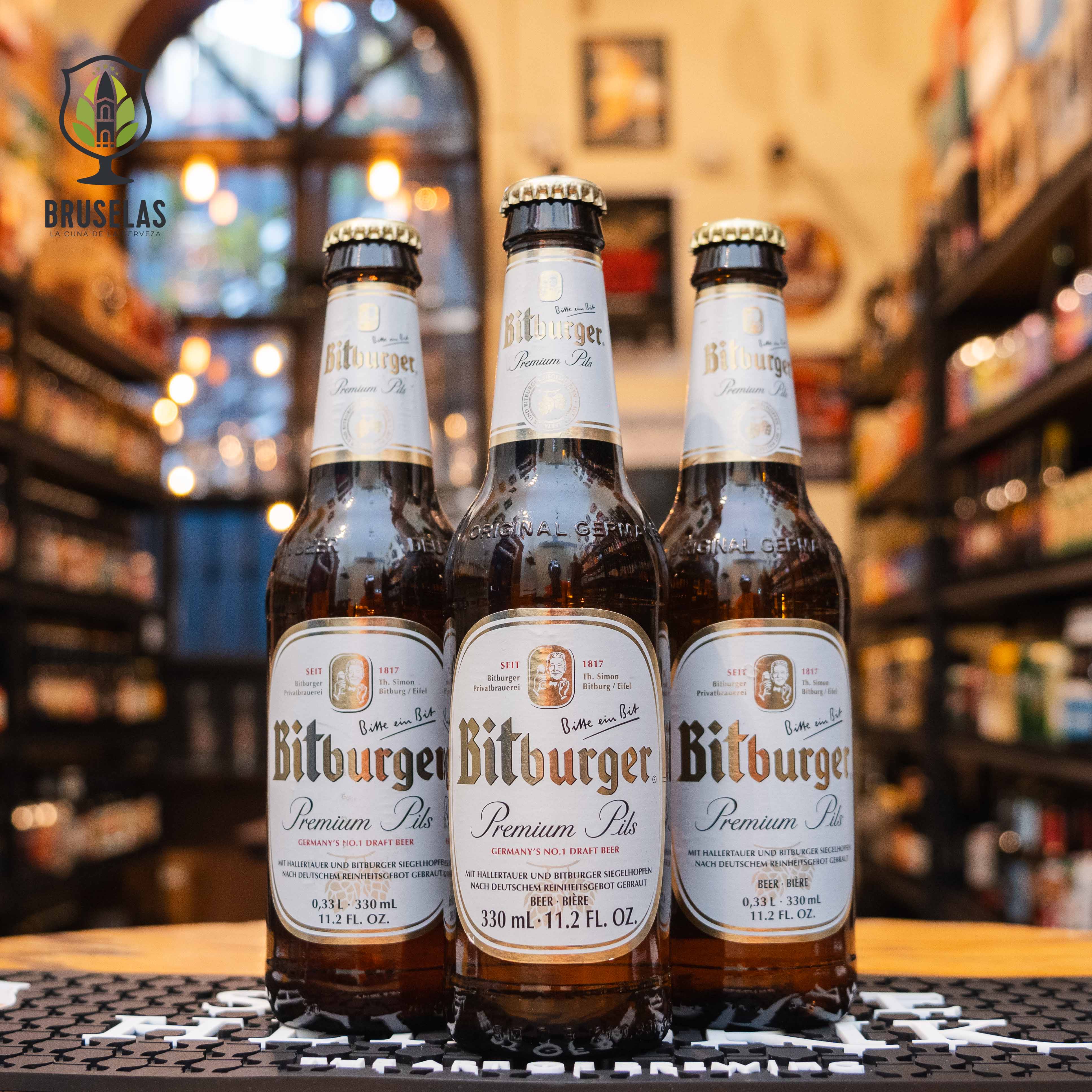 Botella de Bitburger Premium Pils, una pilsner alemana de Bitburger Brauerei. La etiqueta es blanca con detalles dorados y plateados, con el nombre de la marca en letras doradas. ABV de 4.8%, con un sabor refrescante, equilibrado entre notas herbales y un amargor limpio. Ideal para acompañar ensaladas, mariscos y quesos suaves. El diseño clásico de la botella de 330 ml refleja su tradición alemana.