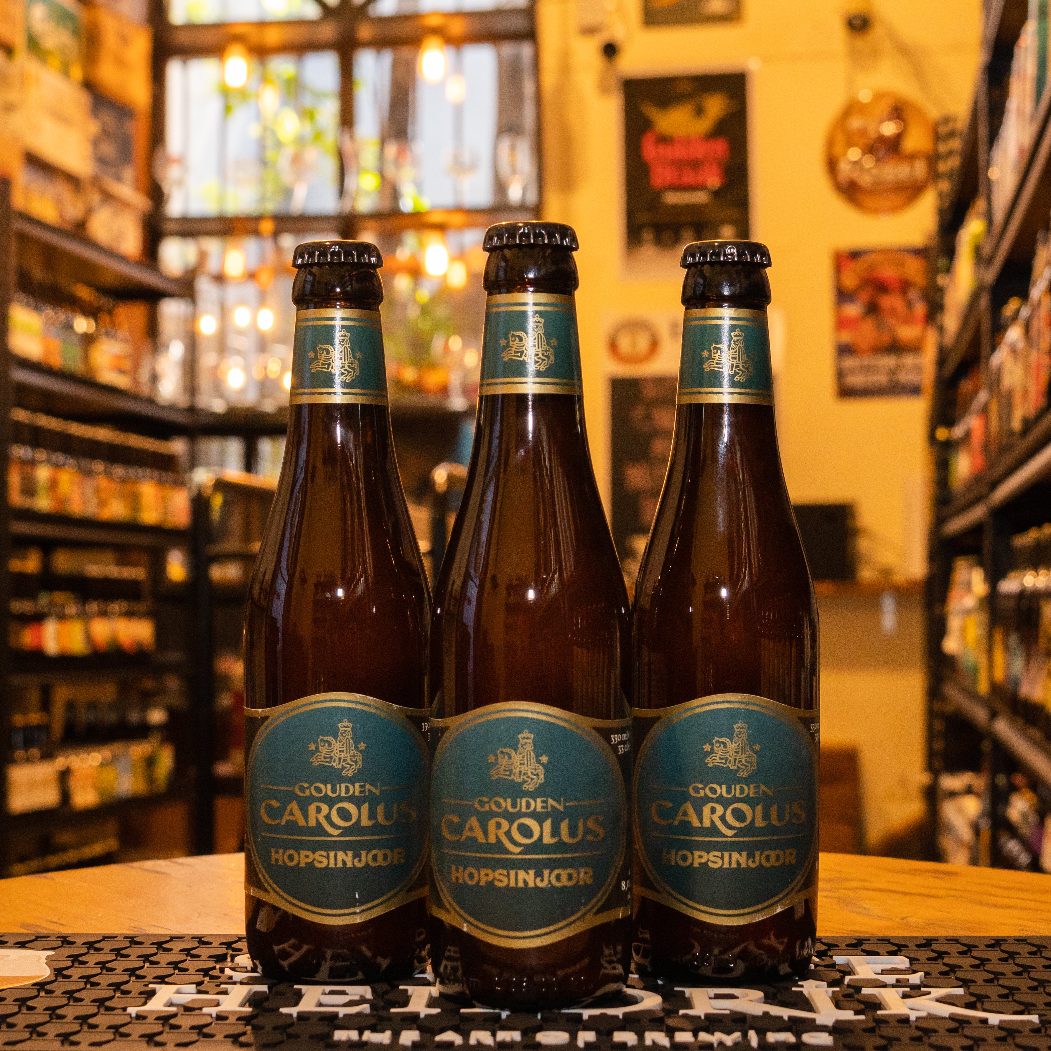 Botella de Gouden Carolus Hopsinjoor, una Belgian Pale Ale de Brouwerij Het Anker. La etiqueta presenta un diseño tradicional en tonos dorados y verdes, destacando su carácter lupulado. Con un ABV de 8.0%, ofrece aromas florales y cítricos con notas de frutas tropicales. Su sabor equilibra la dulzura de la malta y el amargor del lúpulo, con un final seco y refrescante. Ideal para acompañar quesos fuertes, platos de pescado y ensaladas.