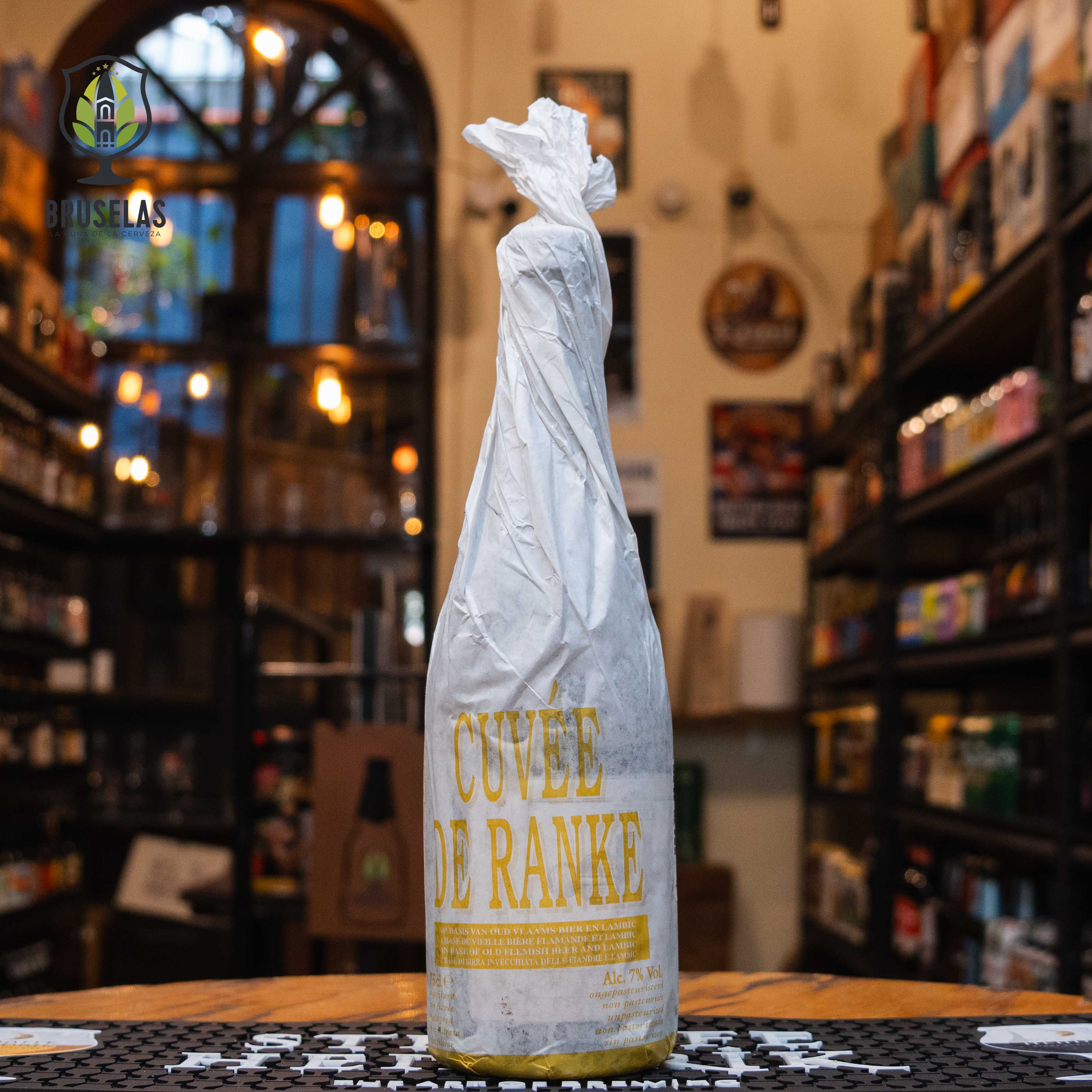 Botella de Cuvée De Ranke, una ale de fermentación mixta de Brouwerij De Ranke, elaborada en Bélgica. La botella está envuelta en un papel blanco con el nombre de la cerveza "Cuvée De Ranke" impreso en letras grandes de color dorado. Presenta un ABV de 7% y ofrece un perfil de sabor ácido, con notas de roble y cítricos. Ideal para acompañar quesos maduros y ensaladas ácidas. El diseño refleja una estética artesanal y rústica.