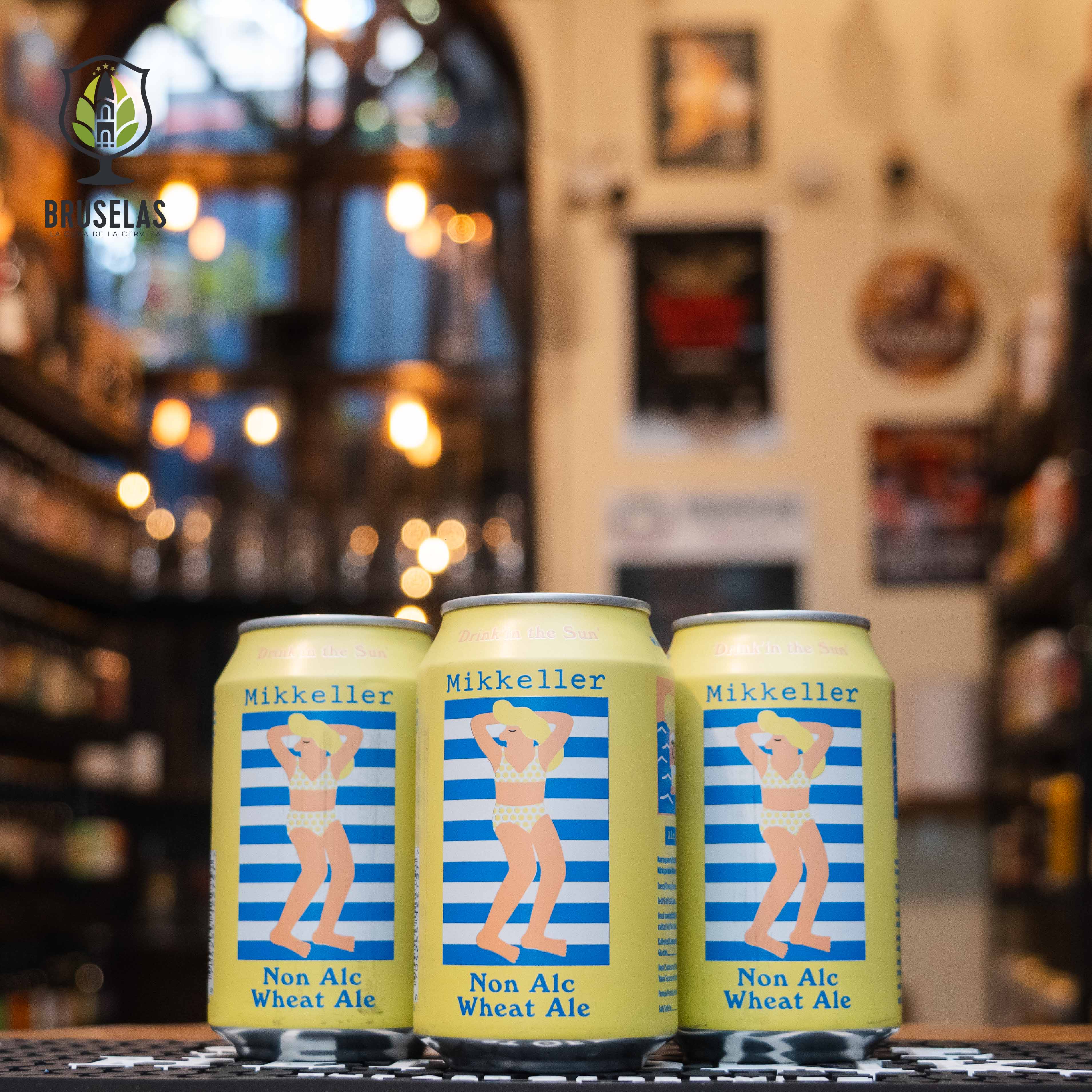 Lata de Mikkeller Drink'in the Sun, una Wheat Ale sin alcohol con un ABV de 0.3%. La etiqueta en tonos amarillos y azules presenta una ilustración de una persona en traje de baño sobre rayas azules. Con aromas cítricos y frutales, y un amargor suave, es ideal para maridar con ensaladas frescas o platillos ligeros de pescado. El diseño refleja la frescura y ligereza de esta cerveza.