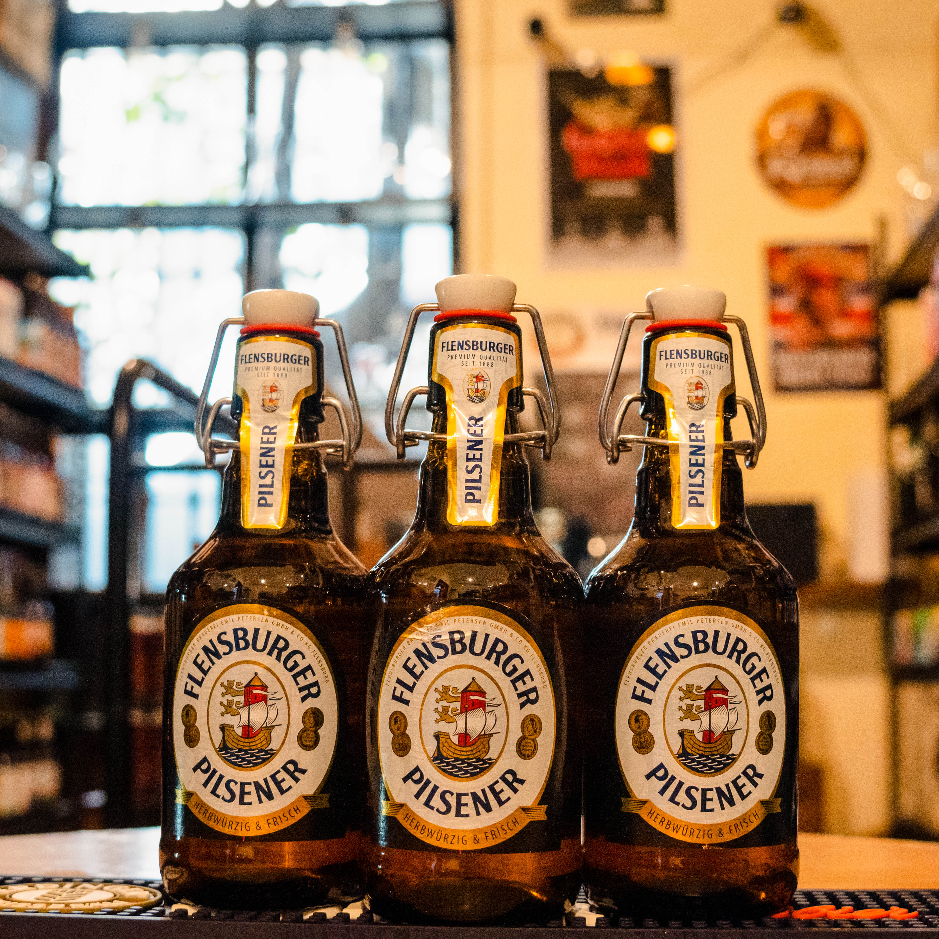 Botella de Flensburger Pilsener, una German Pilsner de Flensburger Brauerei, elaborada en Flensburg, Alemania. La etiqueta presenta tonos verdes y dorados, con el logotipo de la marca y su distintiva tapa de cierre hermético. Con un ABV de 4.8%, ofrece aromas florales y herbales, con un amargor moderado y un final seco. Ideal para maridar con cocina alemana, pescados y quesos con pimienta. Su frescura proviene del uso de agua de manantiales glaciares.