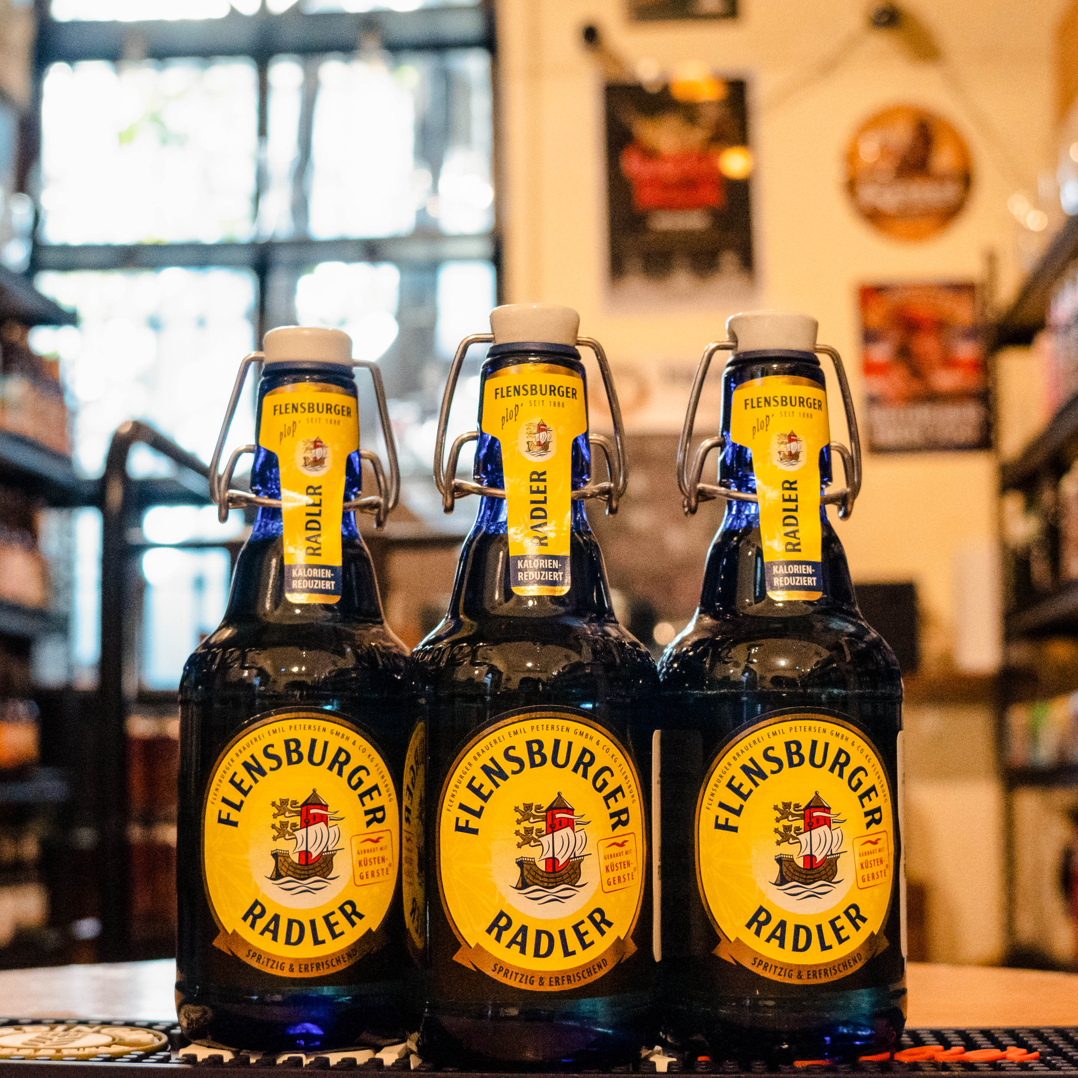 Botella de Flensburger Radler, una mezcla refrescante de cerveza y limonada de la cervecería Flensburger Brauerei, elaborada en Alemania. La etiqueta presenta colores brillantes y cítricos que destacan la frescura del limón. Con un ABV de 2.4%, ofrece un aroma fresco de limón con un sutil toque de malta. Su sabor equilibra la dulzura de la limonada con la ligera amargura de la cerveza, y un final refrescante. Ideal para acompañar ensaladas frescas y pescados ligeros, o disfrutar sola en días calurosos.