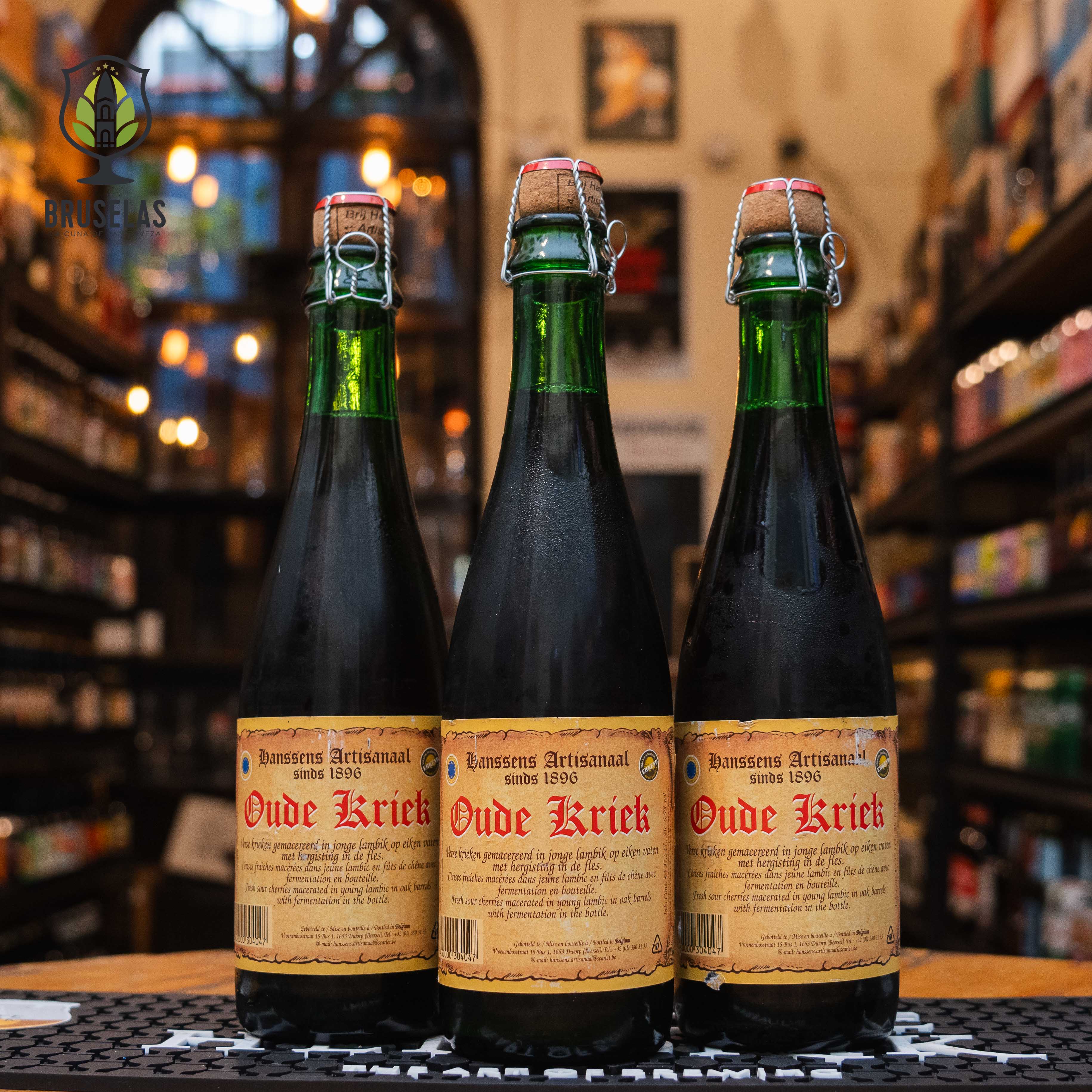 Botella de Oude Kriek, una Lambic de frutas de Hanssens Artisanaal, elaborada en Bélgica. La etiqueta tiene un diseño clásico en tonos beige con letras rojas que destacan el nombre "Oude Kriek". El cierre es de corcho, asegurado con una malla de alambre. Con un ABV de 6%, ofrece un perfil ácido con intensas notas de cereza y un toque de madera. Ideal para acompañar quesos cremosos y postres frutales. El diseño evoca tradición artesanal.