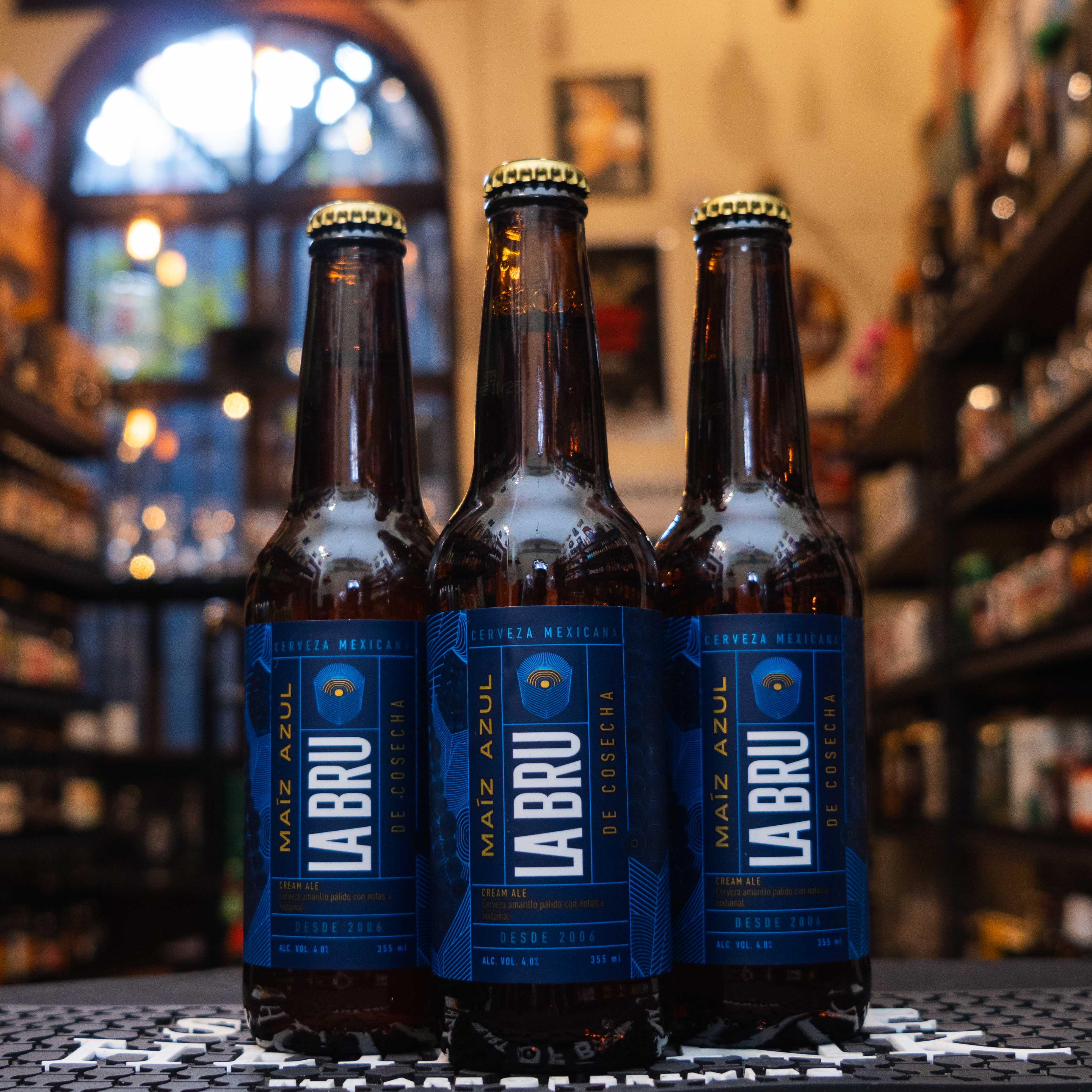 Botella de La Bru Maíz Azul, una Cream Ale de la cervecería mexicana La Bru, elaborada con maíz azul. La etiqueta es azul con detalles dorados y letras blancas en estilo moderno. Con un ABV de 4%, ofrece un sabor ligero, con notas dulces y terrosas del maíz. Ideal para acompañar tacos, tamales y enchiladas. Su diseño destaca la conexión con la gastronomía mexicana.