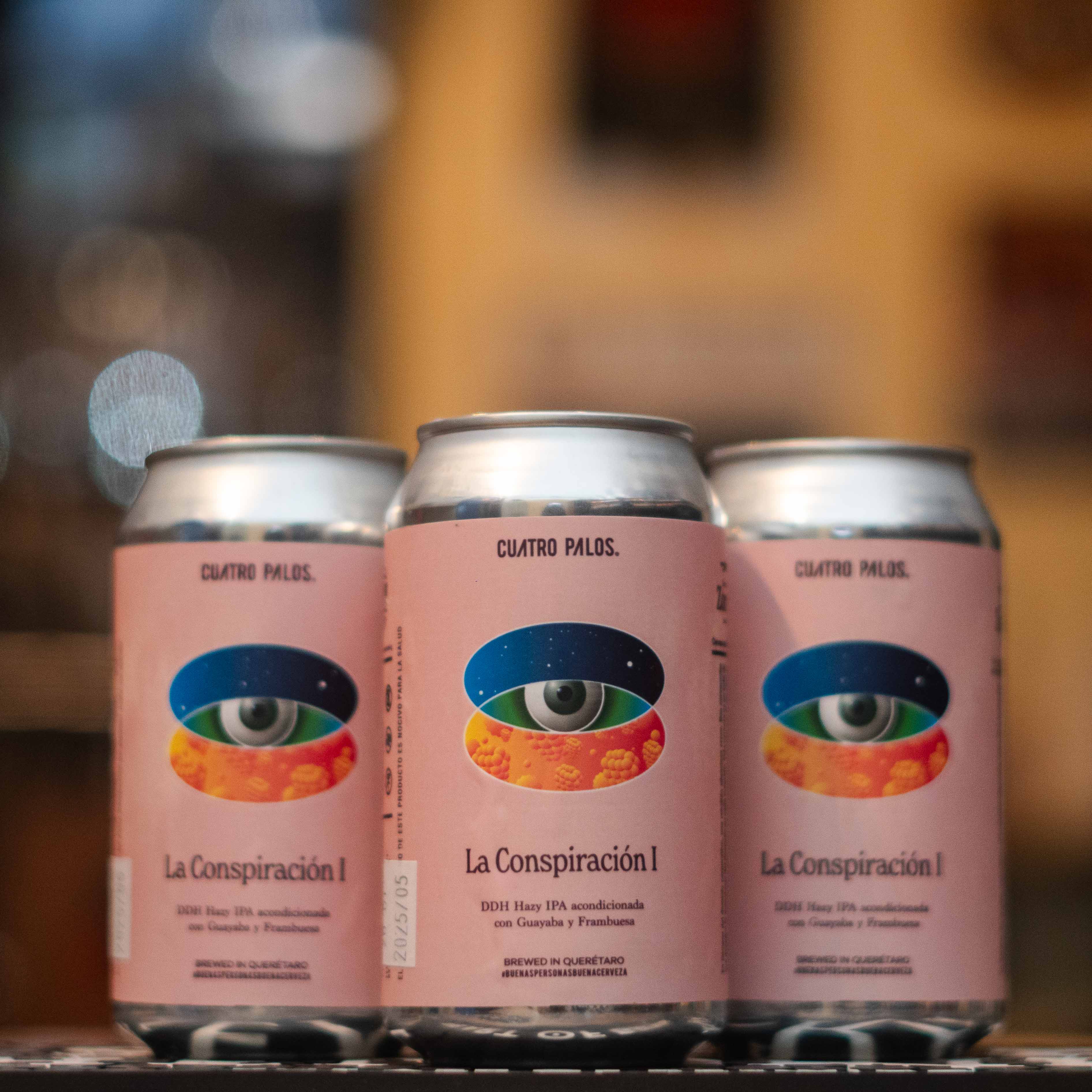 Lata de La Conspiración I, una DDH Hazy IPA de 7% ABV, colaboración entre las cervecerías mexicanas Zorra y Cuatro Palos. La etiqueta rosa destaca un diseño surrealista con un ojo y frutas tropicales. Esta cerveza combina guayaba y frambuesa para ofrecer un perfil afrutado, turbio y cremoso, con notas cítricas del lúpulo. Ideal para maridar con ceviches, ensaladas frescas y postres frutales.