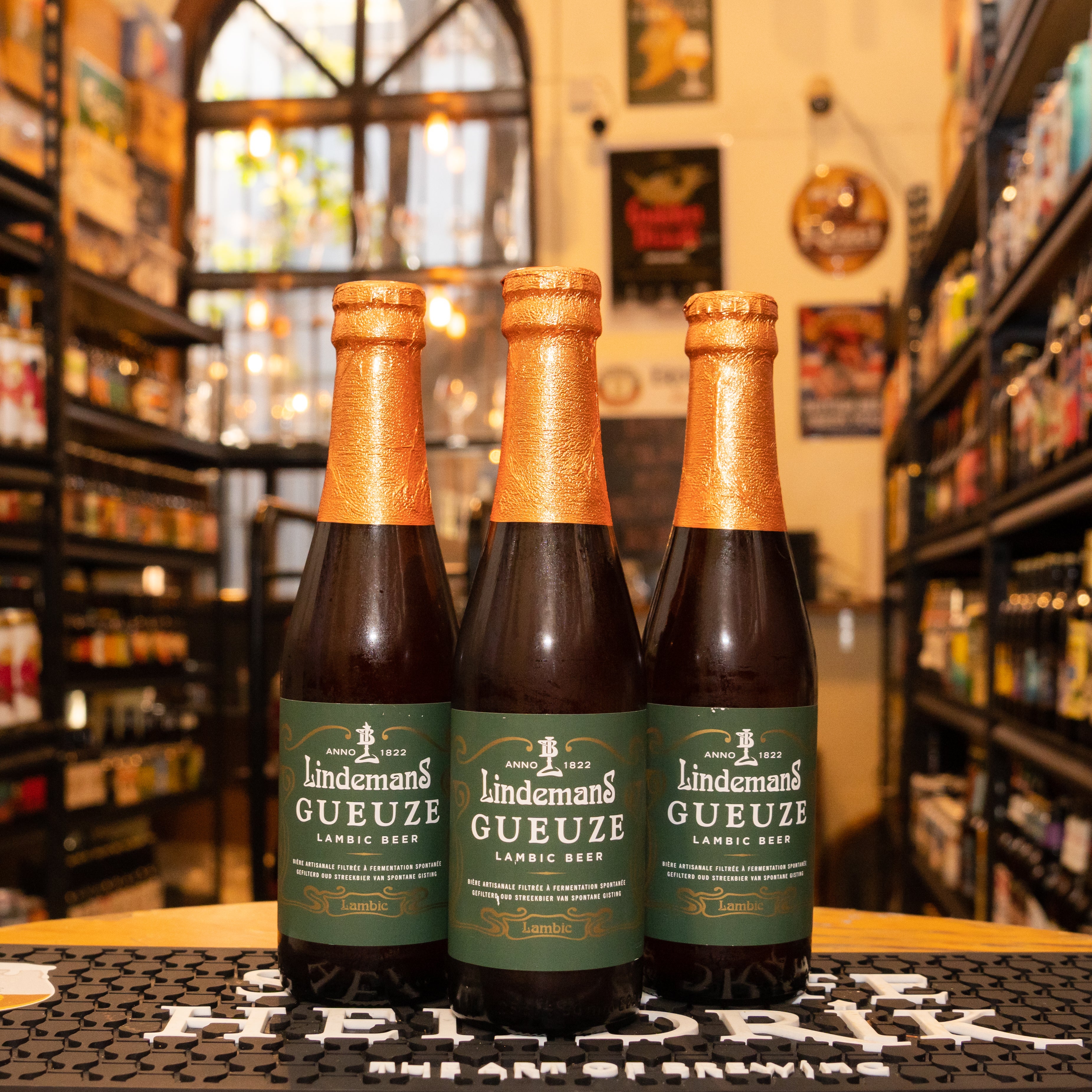 Botella de Lindemans Gueuze, una Gueuze belga de Brouwerij Lindemans. Con un ABV de 5.0%, presenta un color rubio dorado y aromas frescos y afrutados. Su sabor combina dulzura inicial con un acabado seco y ligeramente ácido, con notas que recuerdan al champán y ruibarbo. Ideal para maridar con pescados, mariscos y quesos suaves. El diseño refleja su carácter elegante y refrescante.