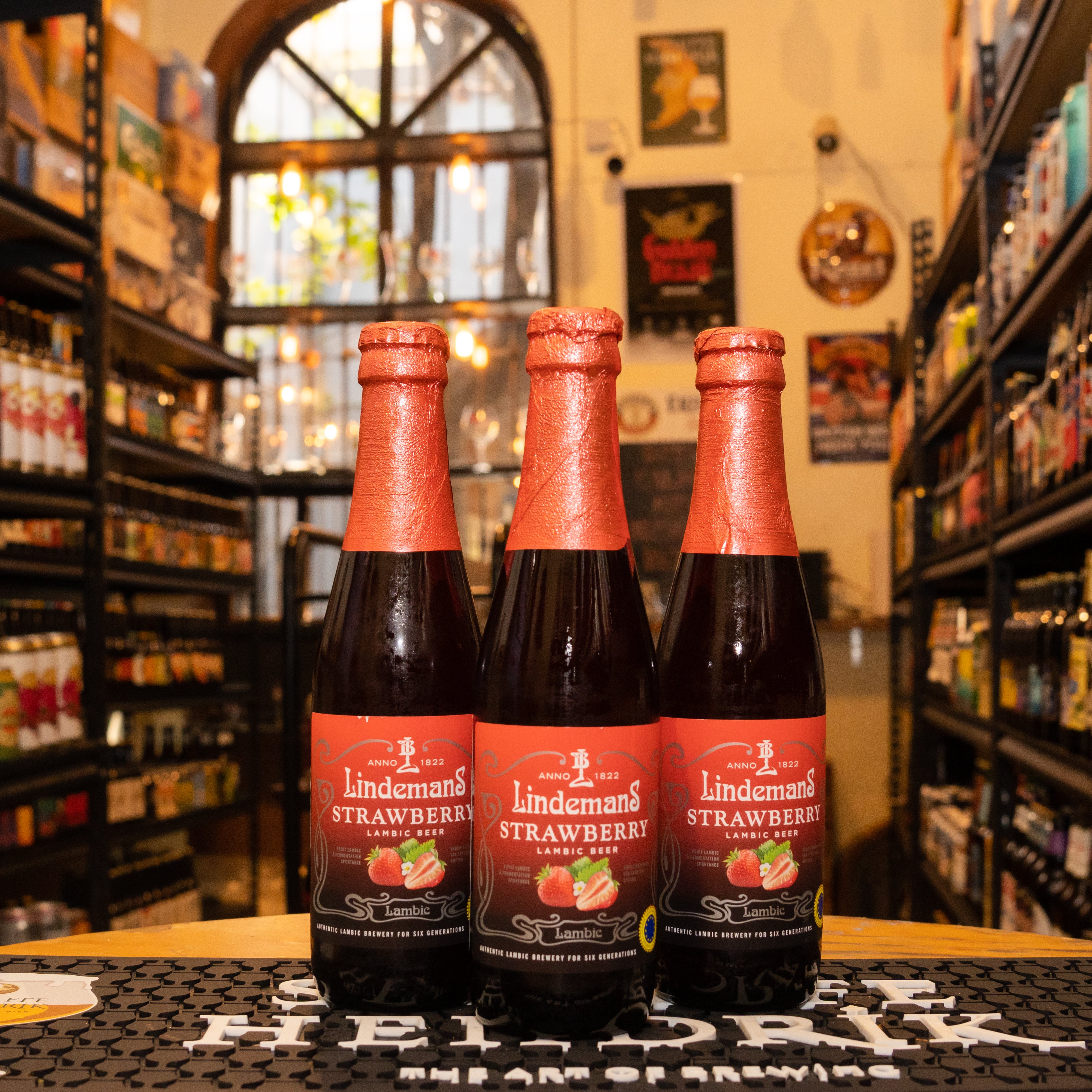 Botella de Lindemans Strawberry, una Fruit Lambic belga de Brouwerij Lindemans. Con un ABV de 4.1%, tiene un color rojo oscuro con matices anaranjados. Elaborada con jugo de fresa y Lambic envejecido en barricas de roble, ofrece un intenso aroma a fresas maduras. Su sabor es afrutado y refrescante, con una acidez equilibrada. Ideal para maridar con quesos suaves, postres como cheesecake y chocolates oscuros.