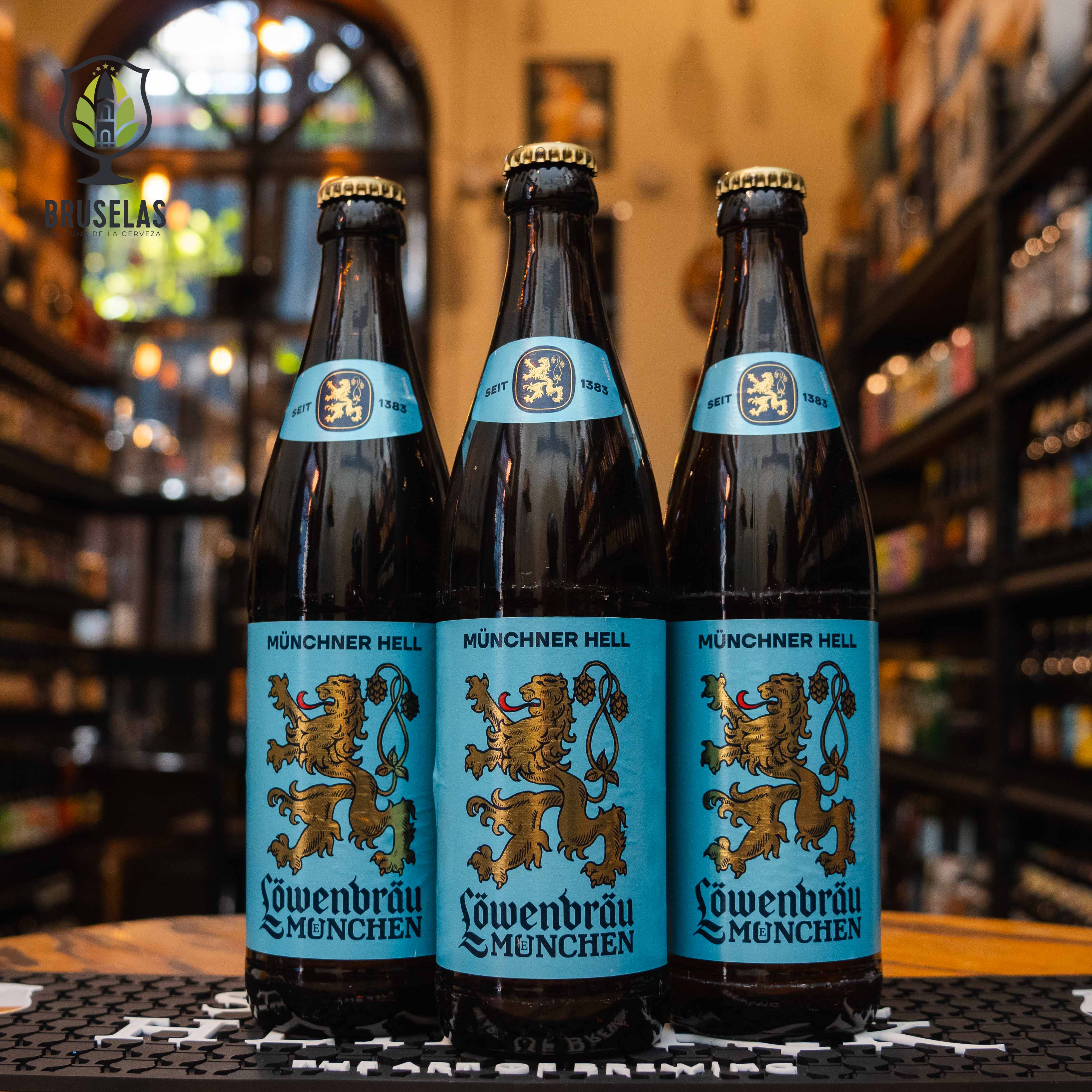 Botella de Löwenbräu Münchner Hell, una cerveza tipo Helles Lager de la cervecería Löwenbräu, elaborada en Múnich, Alemania. La etiqueta azul claro muestra el icónico león bávaro dorado en el centro, junto con el nombre de la marca en letras góticas negras. Con un ABV de 5.2%, esta lager dorada tiene un sabor equilibrado entre la dulzura de la malta y un toque especiado de lúpulo. Ideal para maridar con salchichas bávaras, pretzels y carnes a la parrilla.