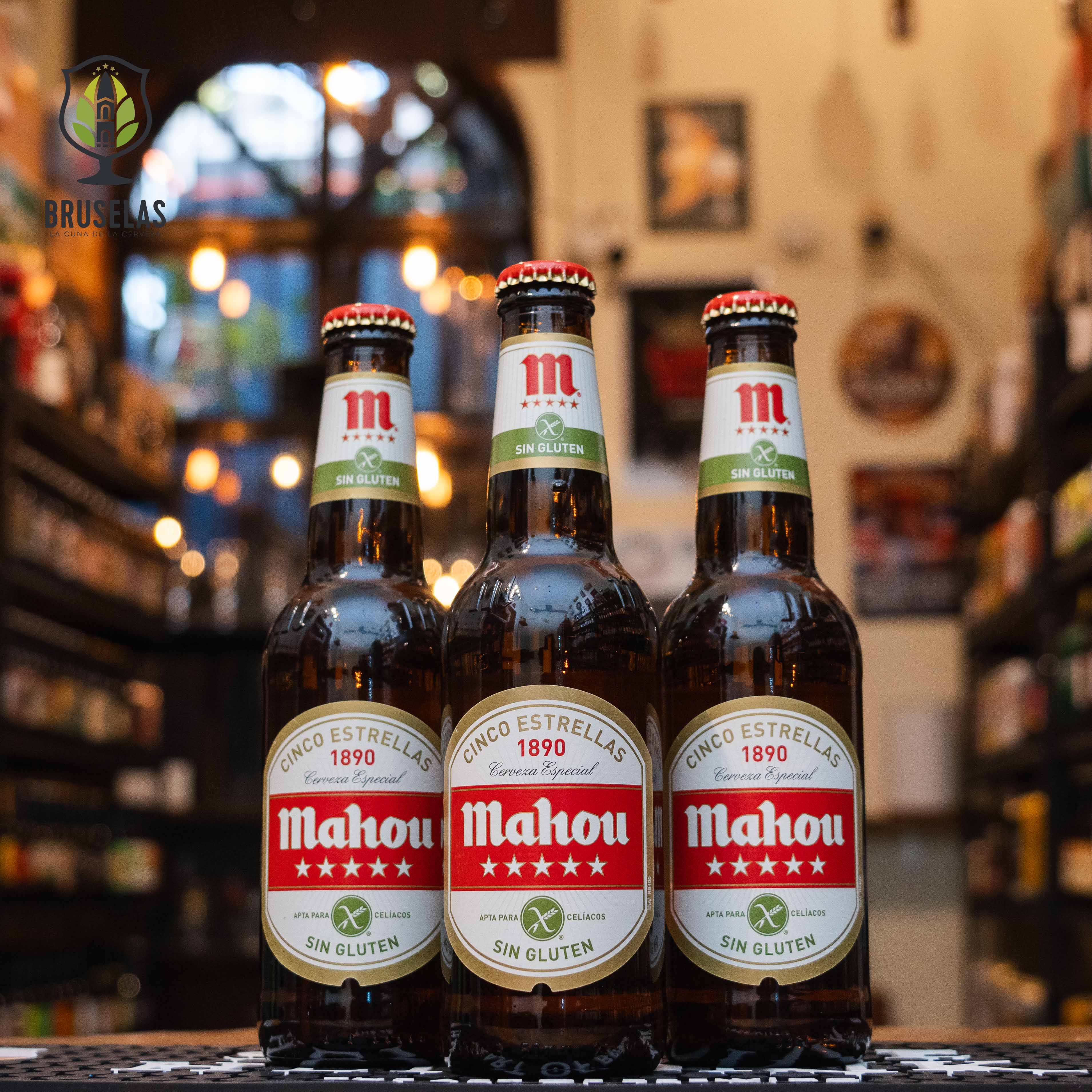 Botella de Mahou Cinco Estrellas Sin Gluten, una lager europea con un ABV de 5.5%. La etiqueta clásica en rojo y blanco destaca su aptitud para celíacos. Con un color dorado brillante y un cuerpo cremoso, ofrece un aroma afrutado y un sabor equilibrado. Ideal para maridar con ensaladas, pescados a la parrilla y tapas españolas. El diseño refleja su autenticidad y calidad tradicional.