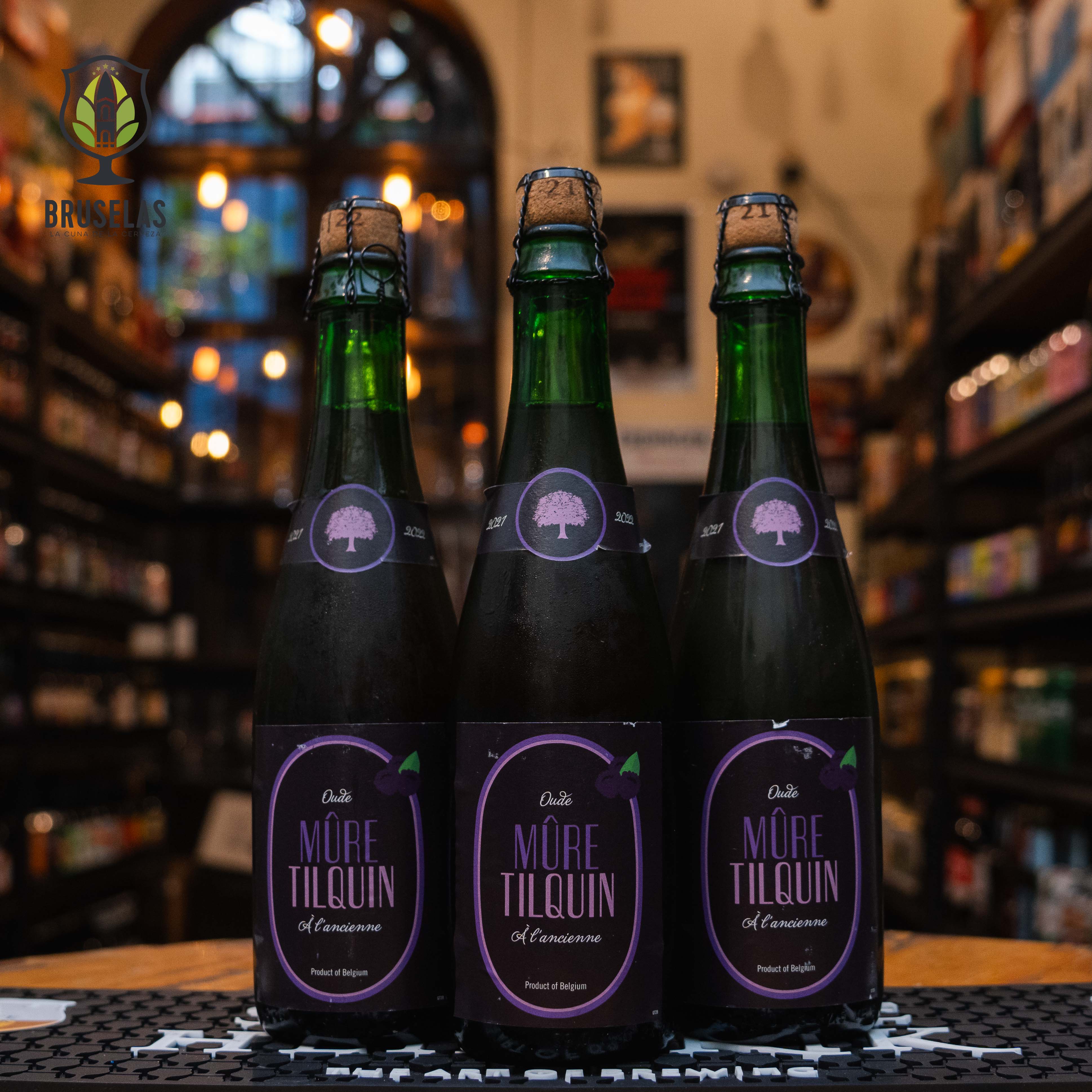 Botella de Tilquin Oude Mûre à l'Ancienne, una lambic de frutas de Gueuzerie Tilquin, elaborada en Bélgica. La etiqueta es predominantemente de color negro y morado, con un diseño minimalista que muestra un árbol en el centro rodeado por un borde circular morado. La tipografía en letras moradas destaca el nombre "Mûre Tilquin". Presenta un ABV de 6.4% y ofrece un perfil de sabor afrutado con un balance entre la acidez del lambic y la dulzura de las moras. Ideal para acompañar quesos fuertes y mariscos.