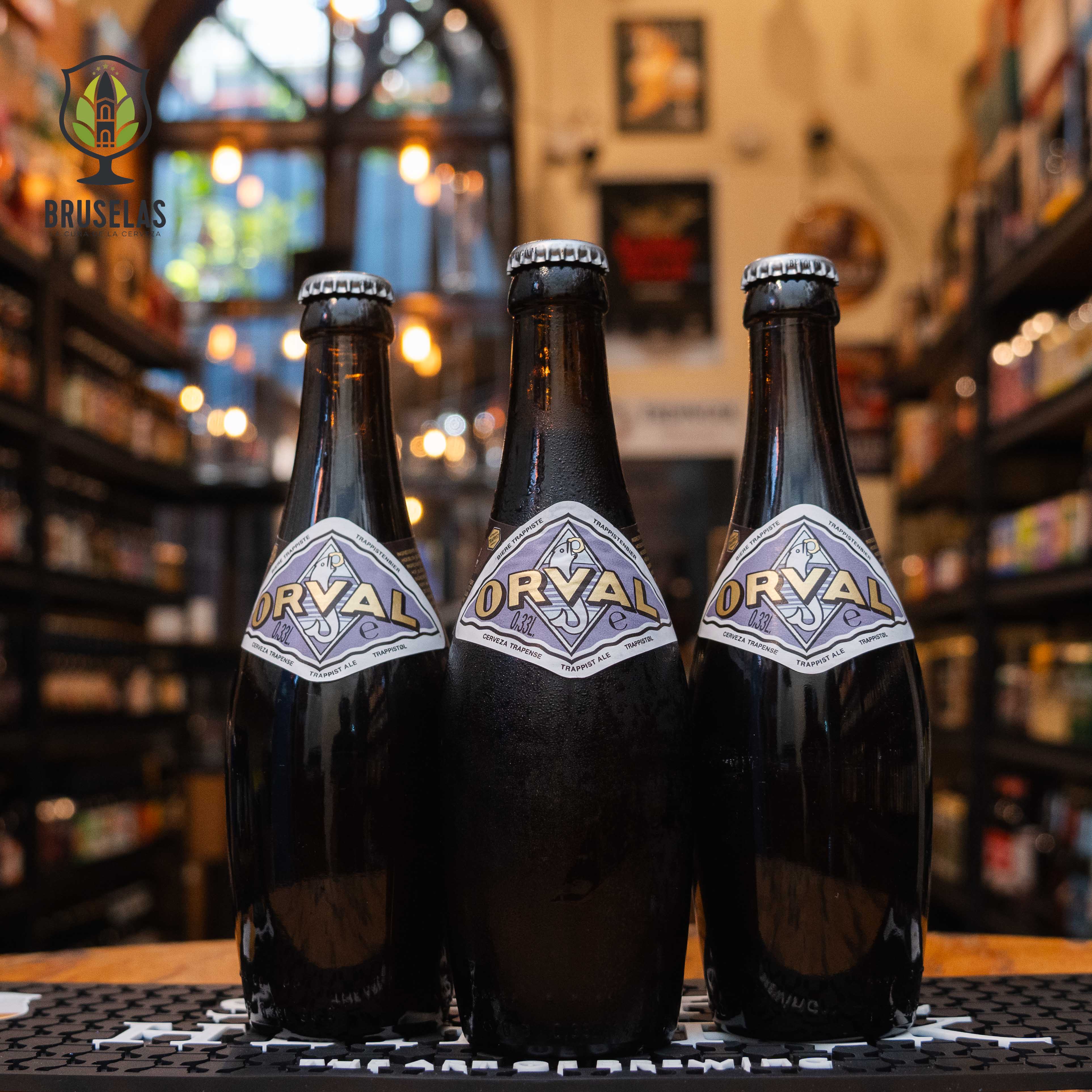 Botella de Orval Trappist Ale, una Belgian Pale Ale elaborada por Brasserie d'Orval en la Abbaye Notre-Dame d'Orval, Bélgica. La etiqueta en forma de rombo tiene tonos lilas y el nombre 'Orval' en letras doradas. Esta cerveza tiene un ABV de 6.9% y ofrece un sabor complejo con notas frutales, un amargor pronunciado y un final seco. Ideal para maridar con quesos, pan moreno, peras y mariscos como mejillones al vapor.