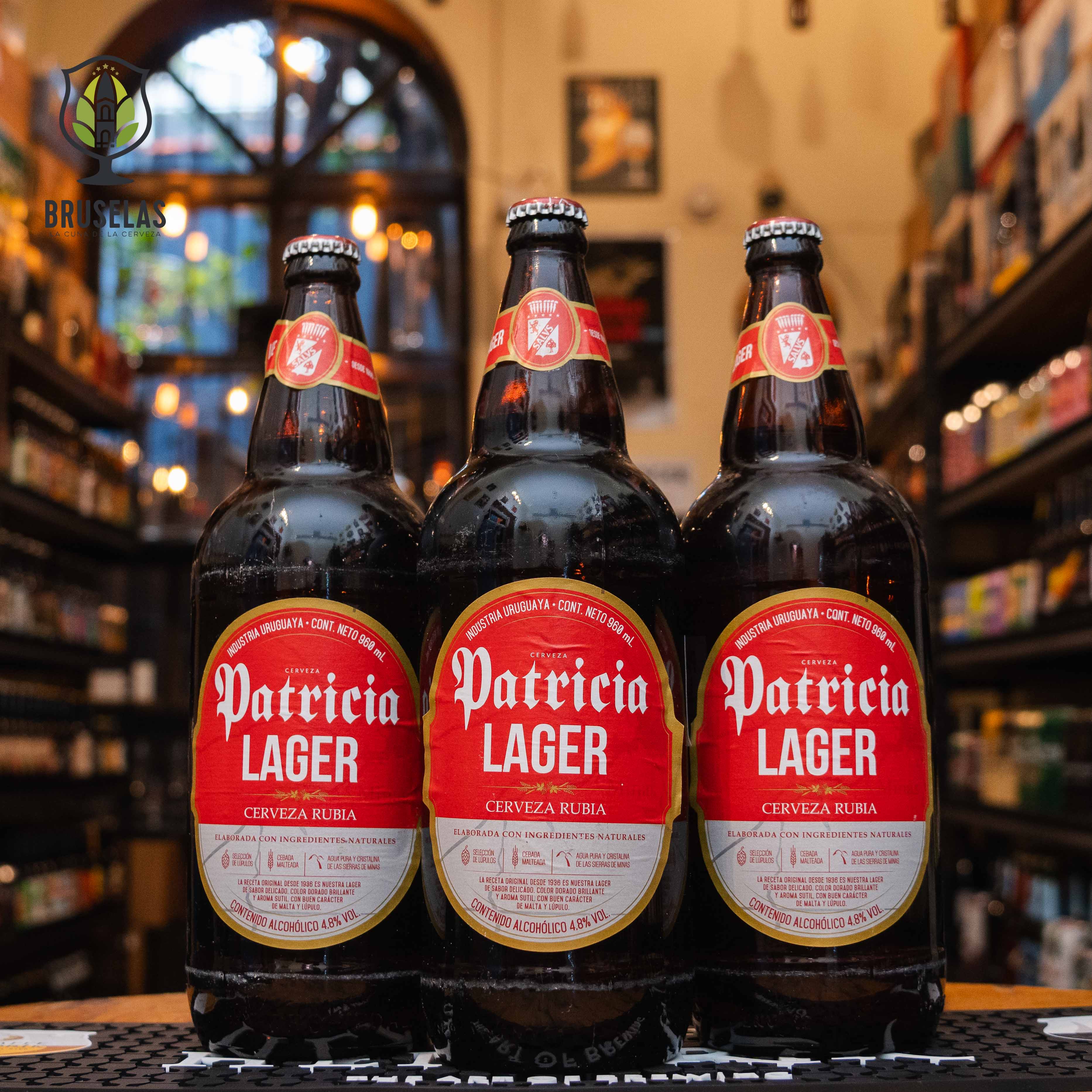 Botella de Patricia Lager, una cerveza lager rubia uruguaya de 960 ml producida por Compañía Salus S.A. La etiqueta presenta un diseño en rojo y blanco, destacando el nombre 'Patricia Lager' en letras blancas. Con un ABV de 4.8%, esta cerveza es suave y refrescante, con notas de malta y un toque herbáceo. Ideal para maridar con asados, empanadas y mariscos, perfecta para celebraciones.