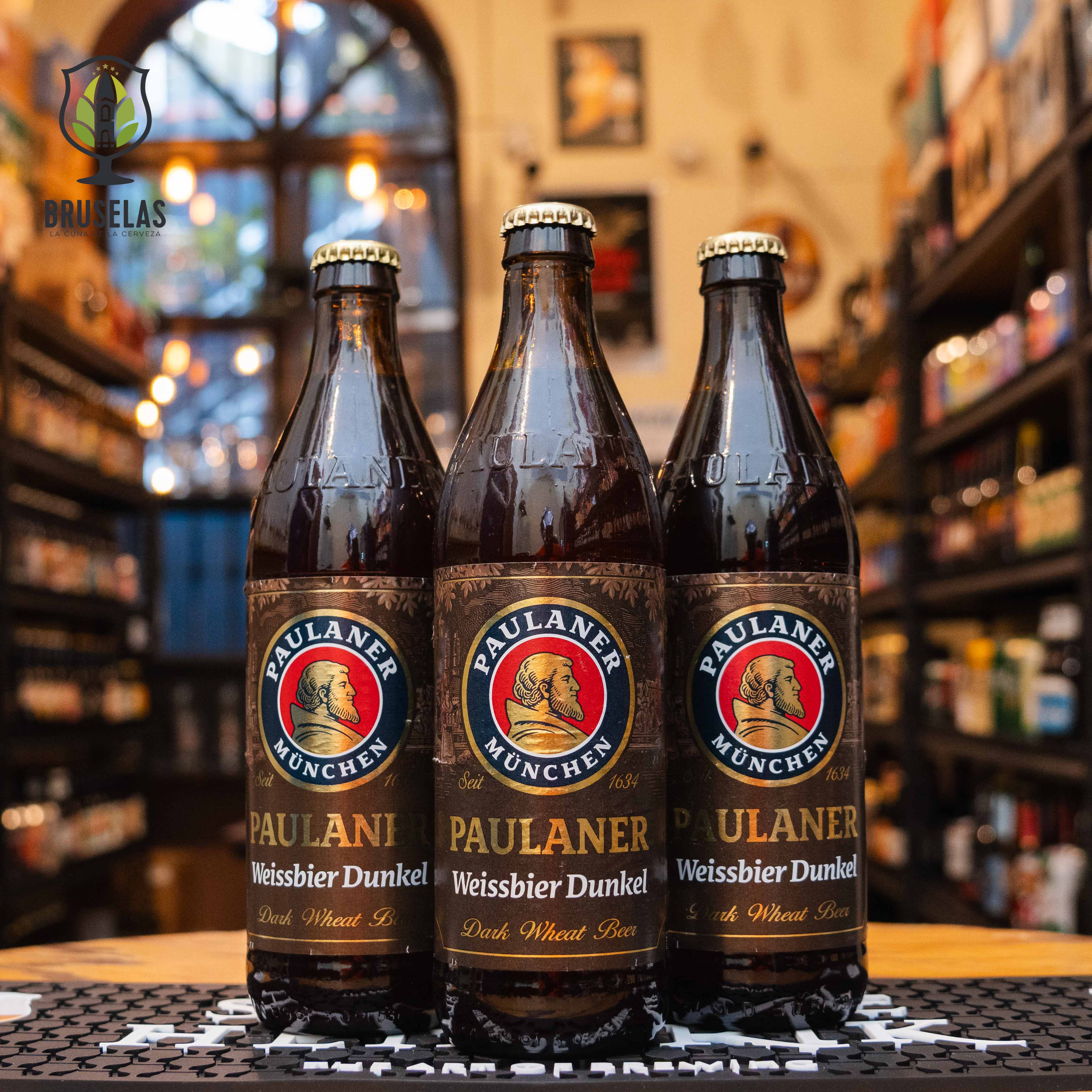 Botella de Paulaner Weissbier Dunkel, una cerveza de trigo oscura elaborada por Paulaner Brauerei en Múnich, Alemania. La etiqueta presenta el icónico logotipo de Paulaner con el monje en el centro, rodeado de tonos marrones y dorados. Con un ABV de 5.3%, esta Dunkelweizen ofrece aromas de chocolate, cacao y frutas tropicales, con sabores a trigo tostado y nuez. Ideal para maridar con guisos de carne y platos bávaros tradicionales.