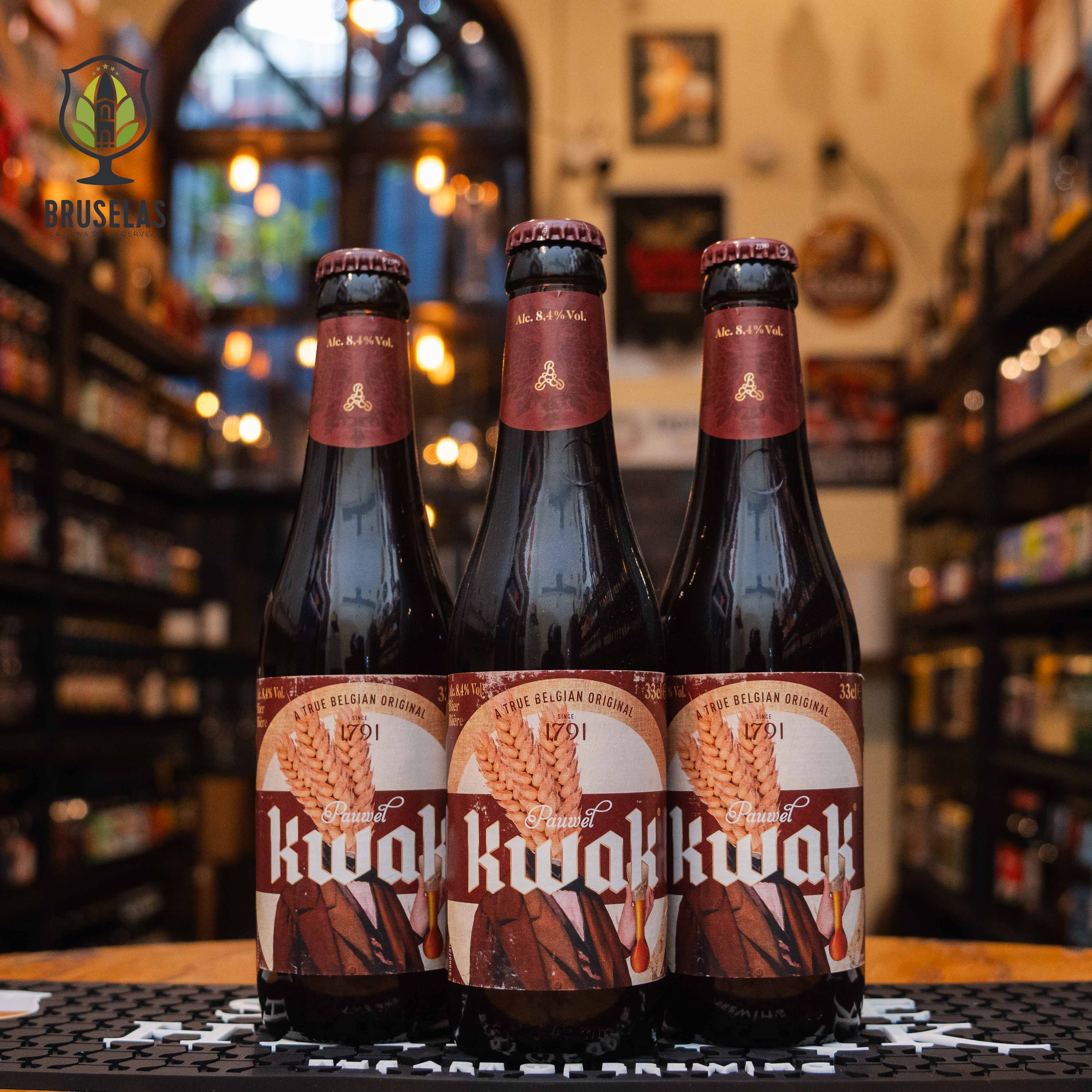 Botella de Pauwel Kwak, una cerveza belga estilo Belgian Strong Ale elaborada por Brouwerij Bosteels, Bélgica. La etiqueta tiene un diseño clásico en tonos marrones, destacando un vaso tradicional de Kwak y detalles dorados de trigo, junto con el nombre "Kwak" en tipografía blanca prominente. Presenta un ABV de 8.4%, con sabores dulces y especiados, y notas de caramelo y frutas tropicales. Ideal para acompañar quesos maduros y carnes asadas.