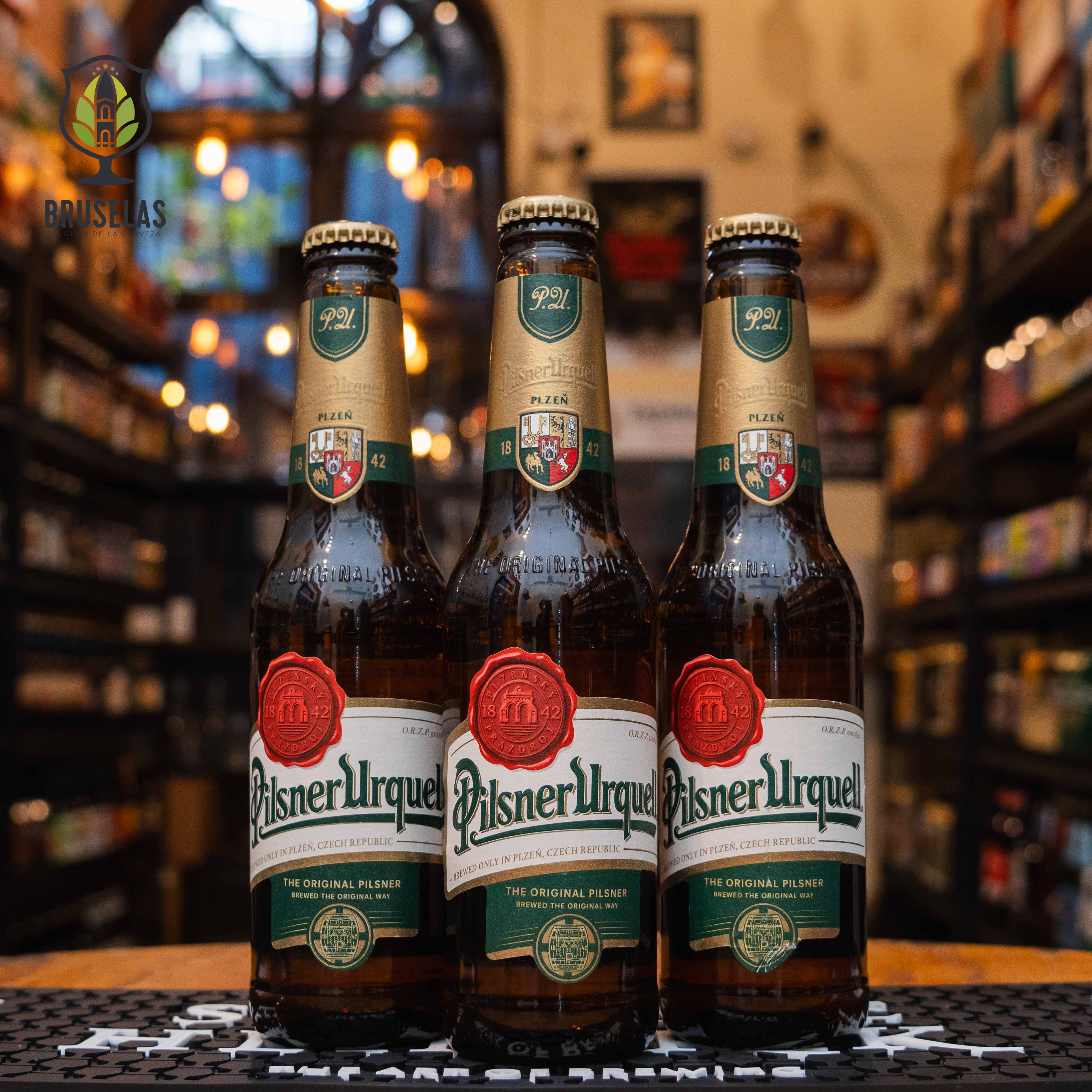 Botella de Pilsner Urquell, una cerveza estilo pilsner elaborada por Pilsner Urquell Brewery en Pilsen, República Checa. La etiqueta presenta un diseño clásico en tonos verdes y dorados, con un sello rojo que resalta su año de origen, 1842. Presenta un ABV de 4.4%, ofreciendo un equilibrio entre dulzura maltosa y el característico amargor del lúpulo Saaz. Ideal para acompañar carnes a la parrilla, mariscos y quesos suaves.