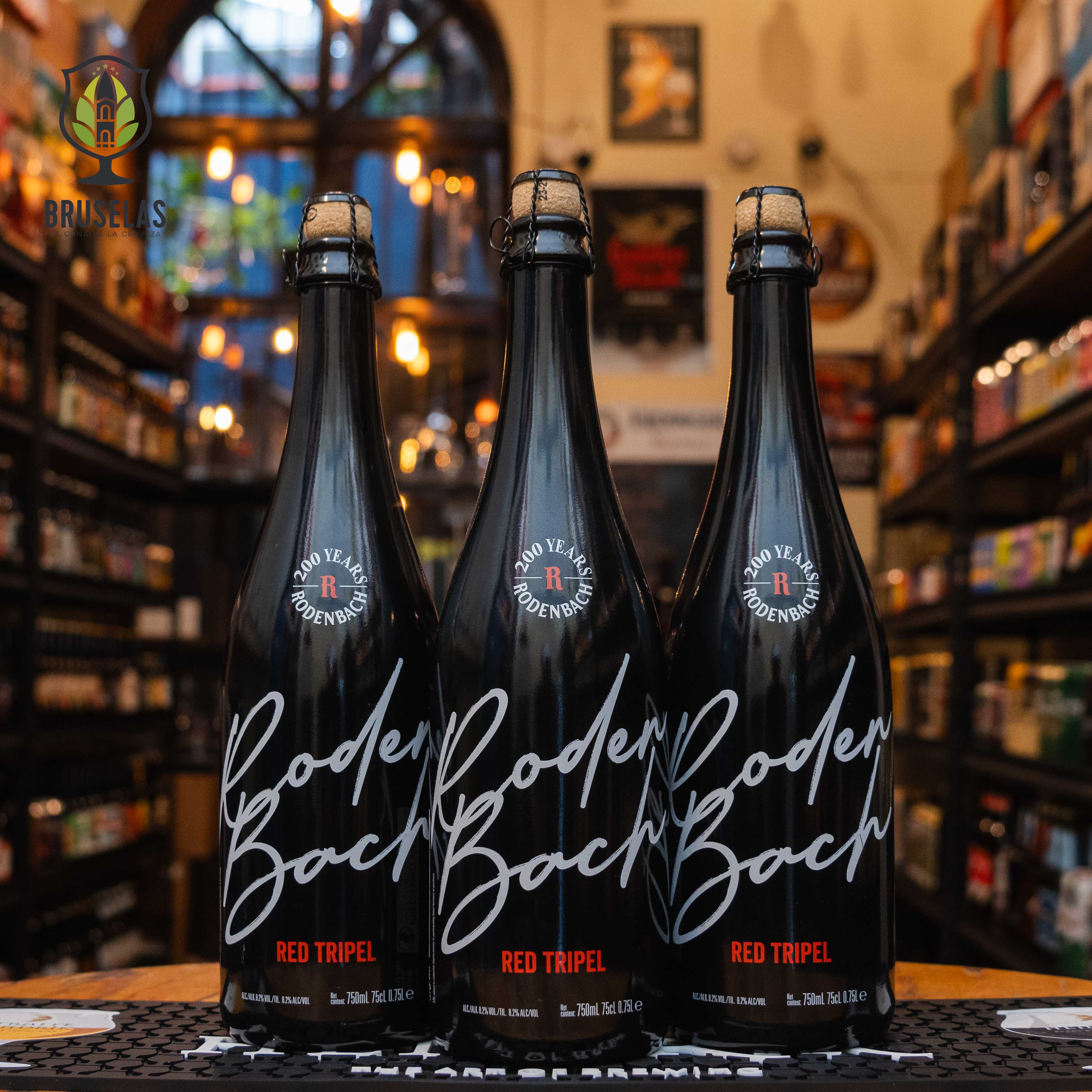 Botella de Rodenbach Red Tripel, una cerveza belga que fusiona Flemish Red Ale y Tripel, elaborada por Rodenbach Brewery. La botella es negra con letras blancas estilizadas y un toque rojo. ABV de 8.5%, con aromas de frutas rojas, malta y especias. Sabor equilibrado con notas ácidas y dulces, ideal para acompañar carnes rojas, estofados y quesos maduros. Diseño moderno y elegante.