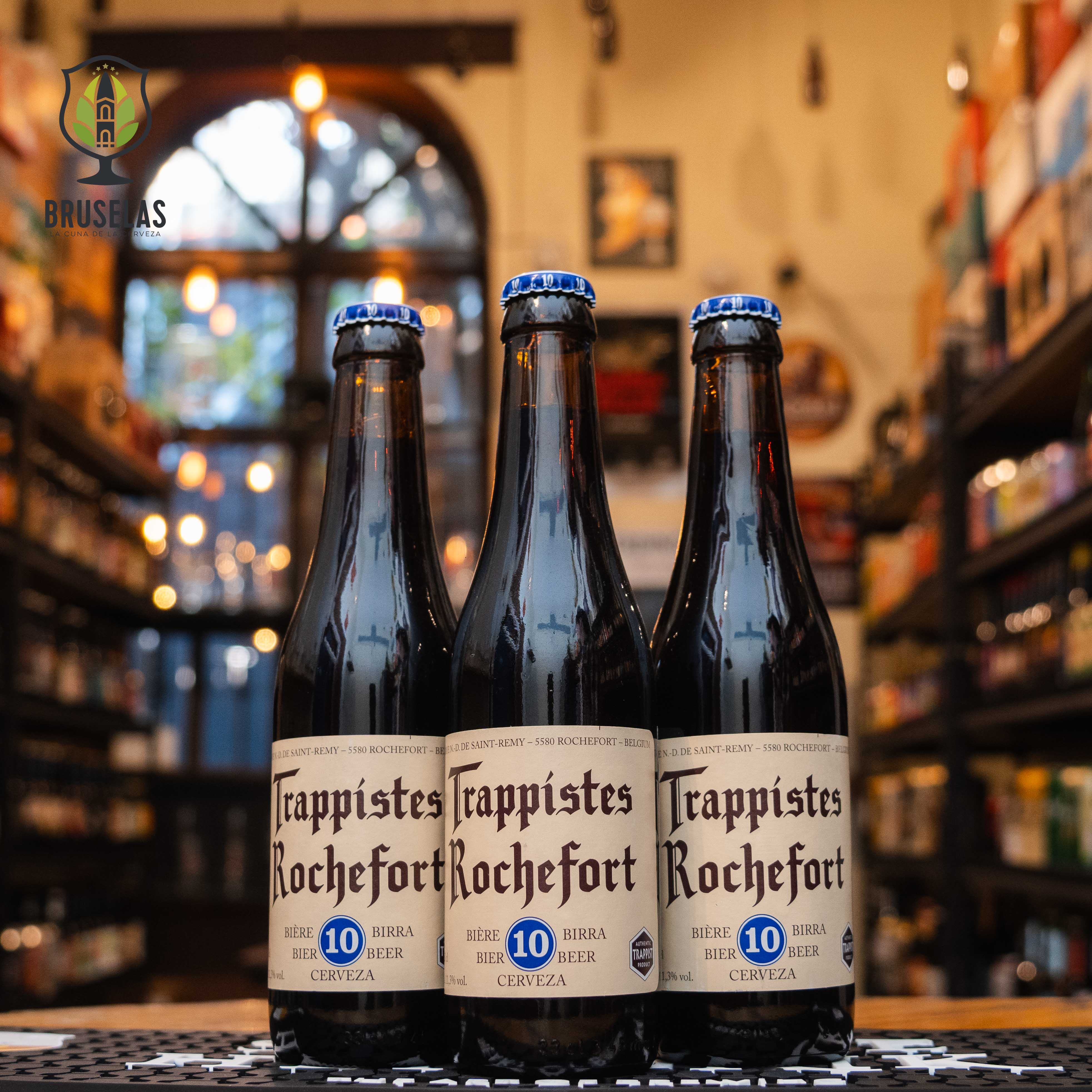 Botella de Trappistes Rochefort 10, una cerveza estilo Quadrupel de la Abadía de Notre-Dame de Saint-Rémy, Bélgica. La etiqueta tiene letras góticas negras sobre fondo beige, con un emblema azul que indica '10'. ABV de 11.3%, con sabores complejos de frutas oscuras, malta dulce y especias. Ideal para acompañar carnes estofadas, quesos fuertes y postres de chocolate. Diseño tradicional trapense.