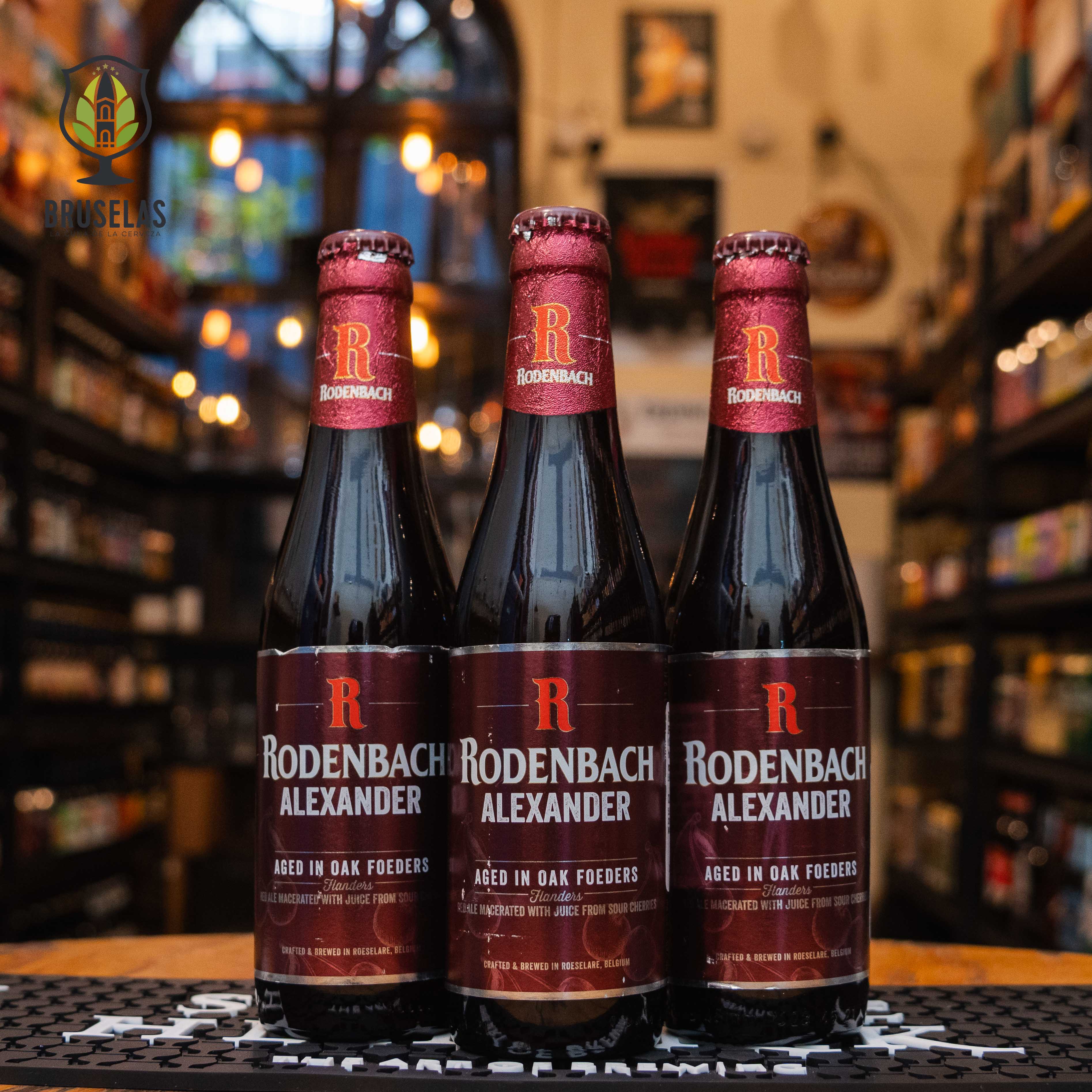 Botella de Rodenbach Alexander, una Flanders Red Ale de Brouwerij Rodenbach, Bélgica. La etiqueta presenta un fondo rojo Botella de Rodenbach Alexander, una Flanders Red Ale de Brouwerij Rodenbach, Bélgica. La etiqueta presenta un fondo rojo oscuro con una 'R' en grande y letras blancas. ABV de 5.6%, con aromas y sabores de cerezas ácidas, madera y un toque de vainilla. Ideal para acompañar quesos de cabra o azul, carnes a la parrilla y postres de chocolate. El diseño refleja la tradición belga en cervezas 