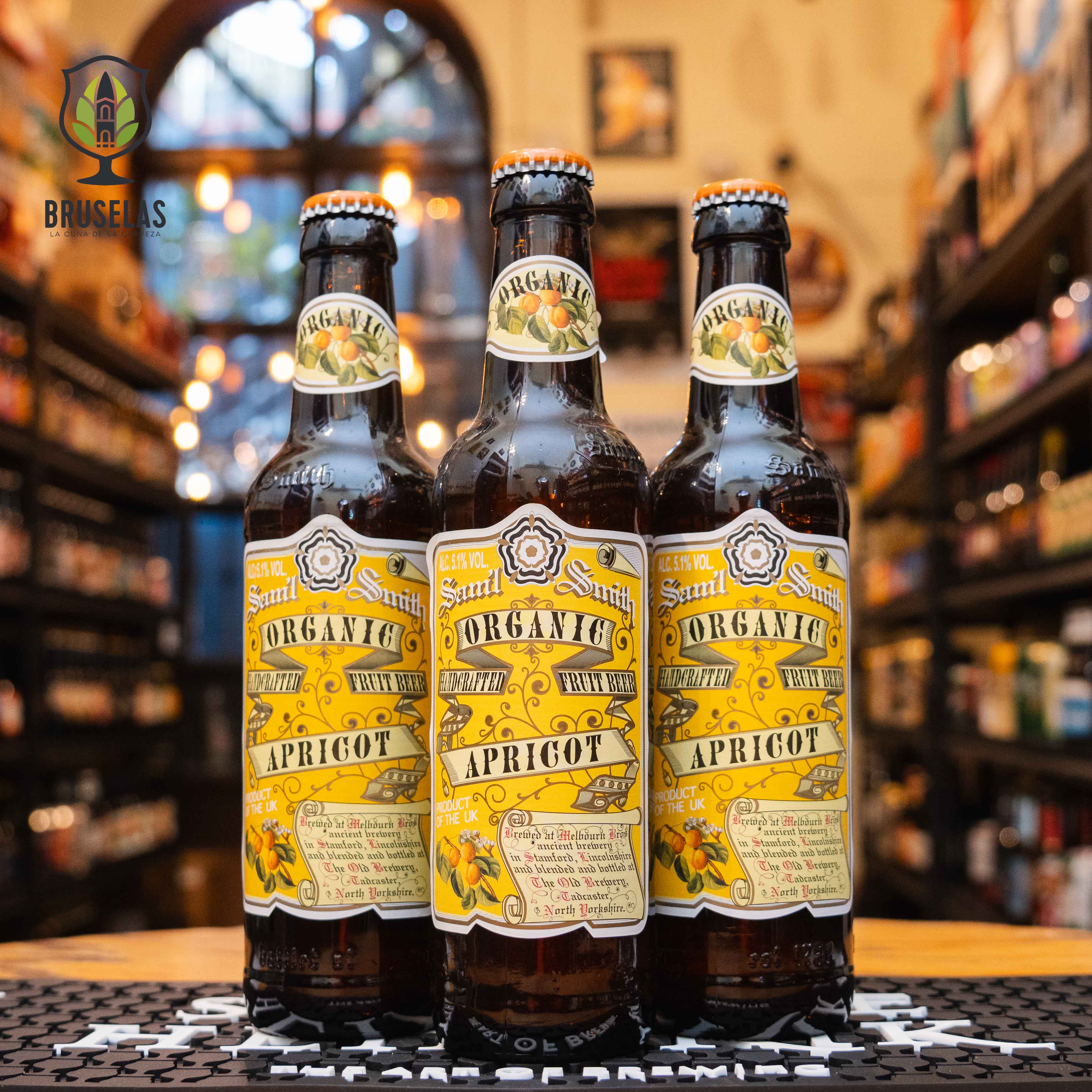 Botella de Samuel Smith Organic Apricot, una Fruit Beer elaborada en Inglaterra por Samuel Smith Old Brewery. La etiqueta es amarilla con detalles frutales de albaricoque y letras clásicas. ABV de 5.1%, con aromas de albaricoques frescos y malta suave. Sabor dulce y refrescante. Ideal para acompañar tartas de frutas, quesos suaves y ensaladas. Diseño artesanal y orgánico.