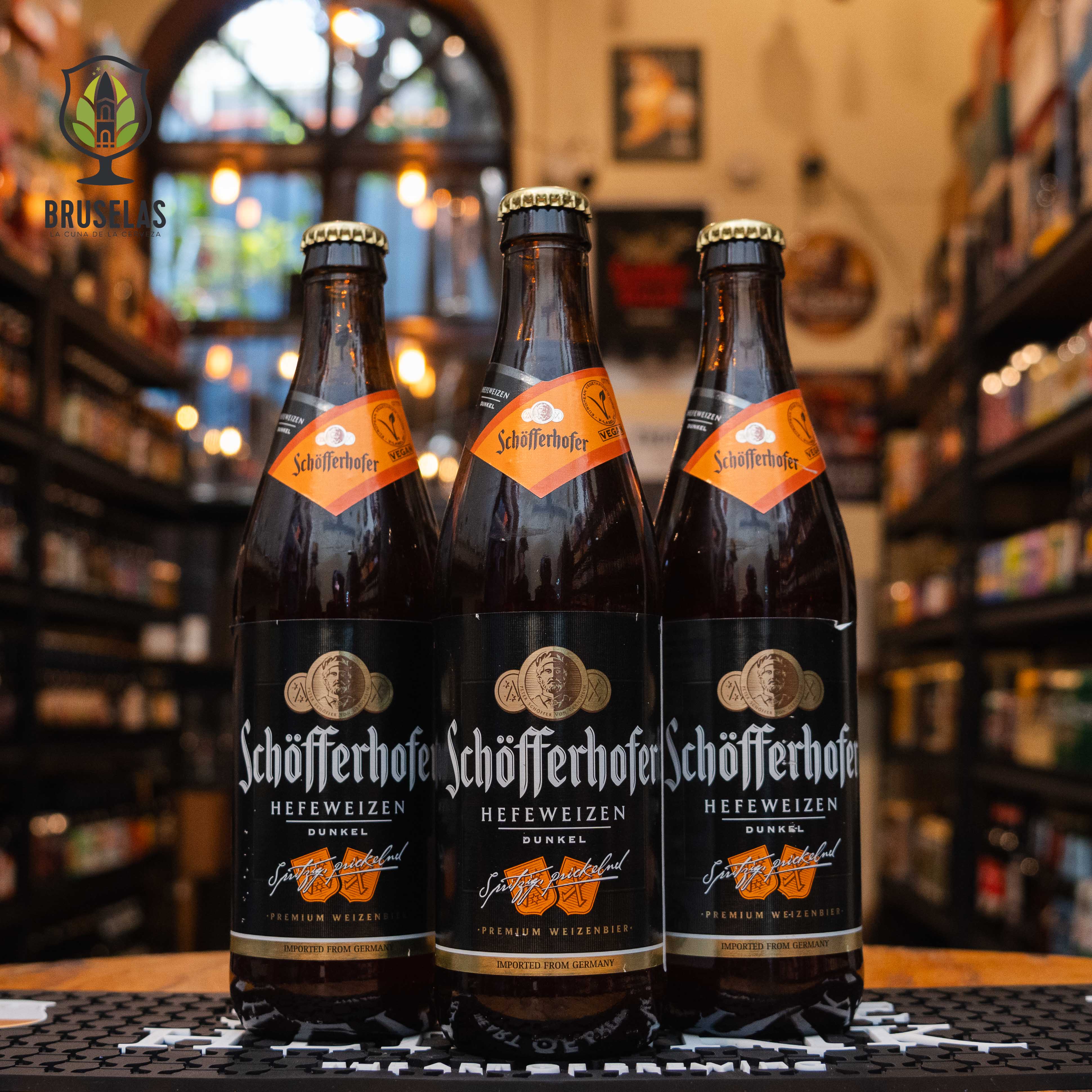 Botella de Schöfferhofer Dunkel, una Dunkelweizen alemana elaborada por Schöfferhofer (Binding-Brauerei). La etiqueta es negra y naranja, con detalles dorados. ABV de 5.0%, con aromas especiados de clavo y canela, y notas de frutas maduras. Sabor dulce con un final tostado y ligeramente especiado. Ideal para acompañar carnes asadas, platos especiados y postres de chocolate. Diseño elegante.