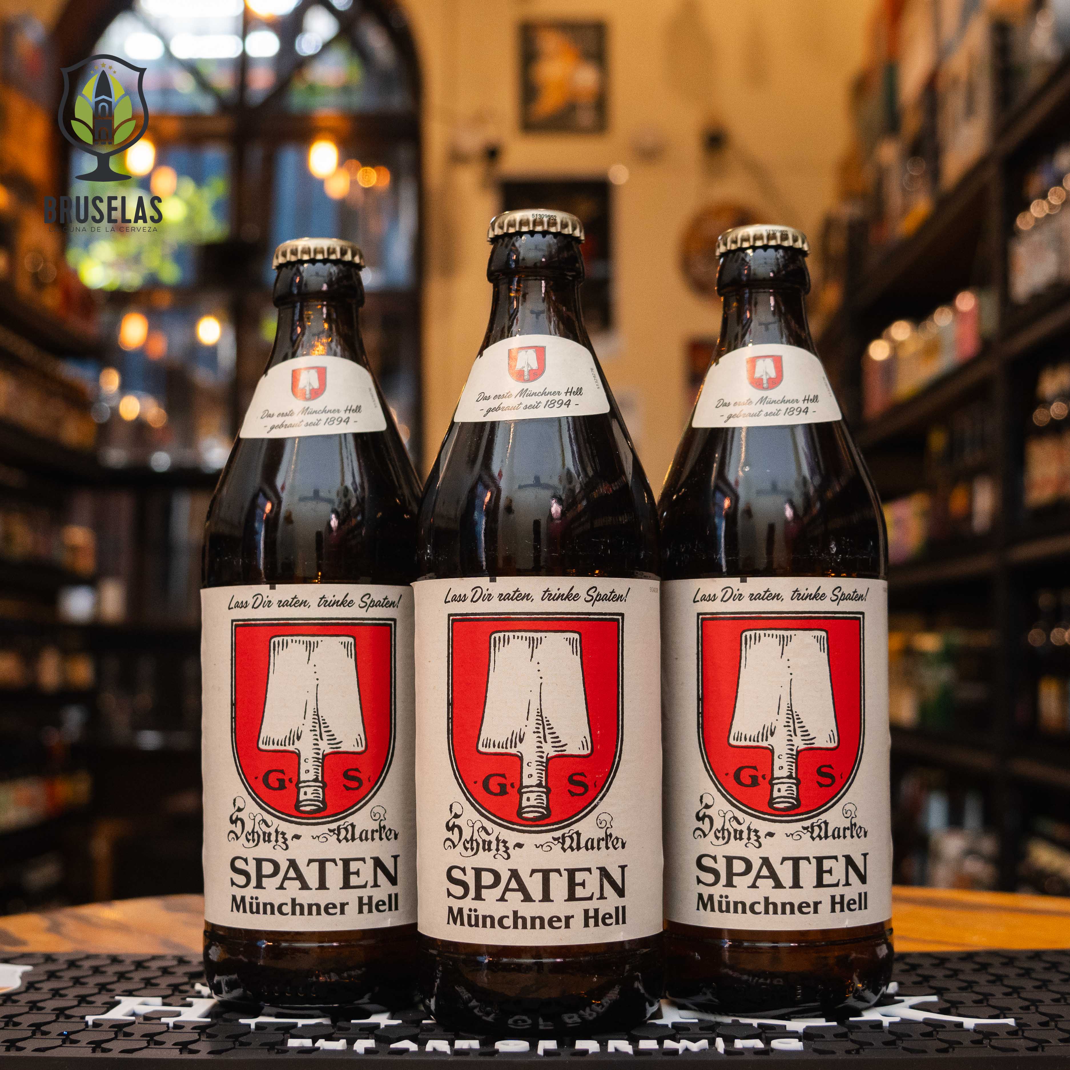 Botella de Spaten Münchner Hell, una Helles lager de la cervecería Spaten-Franziskaner-Bräu en Múnich, Alemania. La etiqueta presenta un escudo rojo con un barril. ABV de 5.2%, con un aroma maltoso y toques de lúpulo suave. Sabor equilibrado con un final ligeramente amargo. Ideal para acompañar carnes blancas, ensaladas y quesos suaves. Lager fresca y fácil de beber.