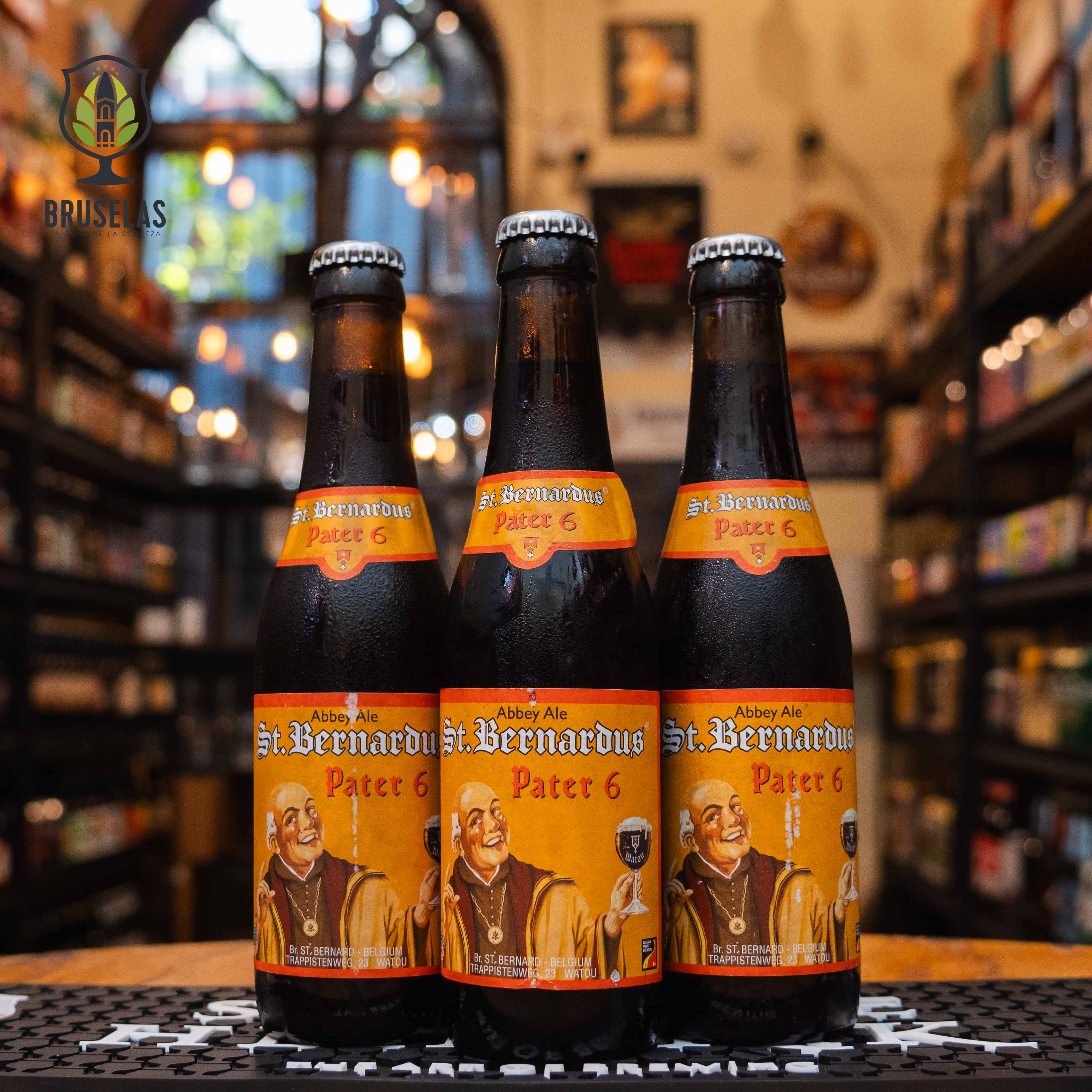 Botella de St. Bernardus Pater 6, una Dubbel belga de la cervecería Brouwerij St. Bernardus, con un ABV de 6.7%. La etiqueta muestra a un monje sonriente con fondo naranja, sosteniendo una copa de cerveza. De color marrón oscuro con espuma densa, presenta aromas de caramelo, frutas oscuras y especias. Su sabor suave incluye notas de malta tostada, higos y pasas, ideal para maridar con guisos de carne y quesos semicurados.