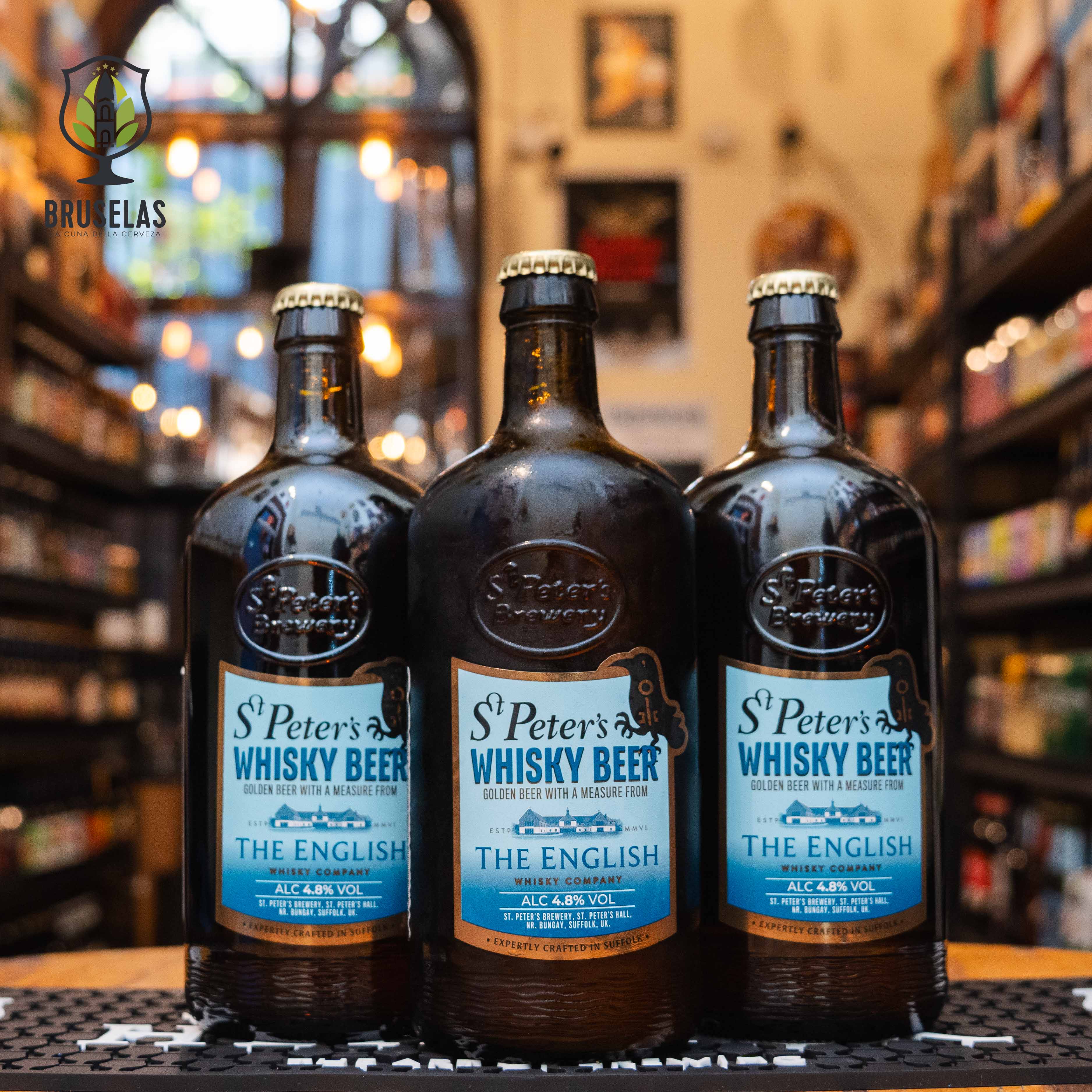 Botella de St. Peter's Whisky Beer, una cerveza dorada con whisky de St. Peter's Brewery y The English Whisky Company. La etiqueta es azul con letras blancas y detalles en marrón. ABV de 4.8%. Ofrece un sabor ahumado de malta de turba con toques maltosos. Ideal para carnes ahumadas y quesos fuertes. La botella tiene un diseño tradicional en relieve.