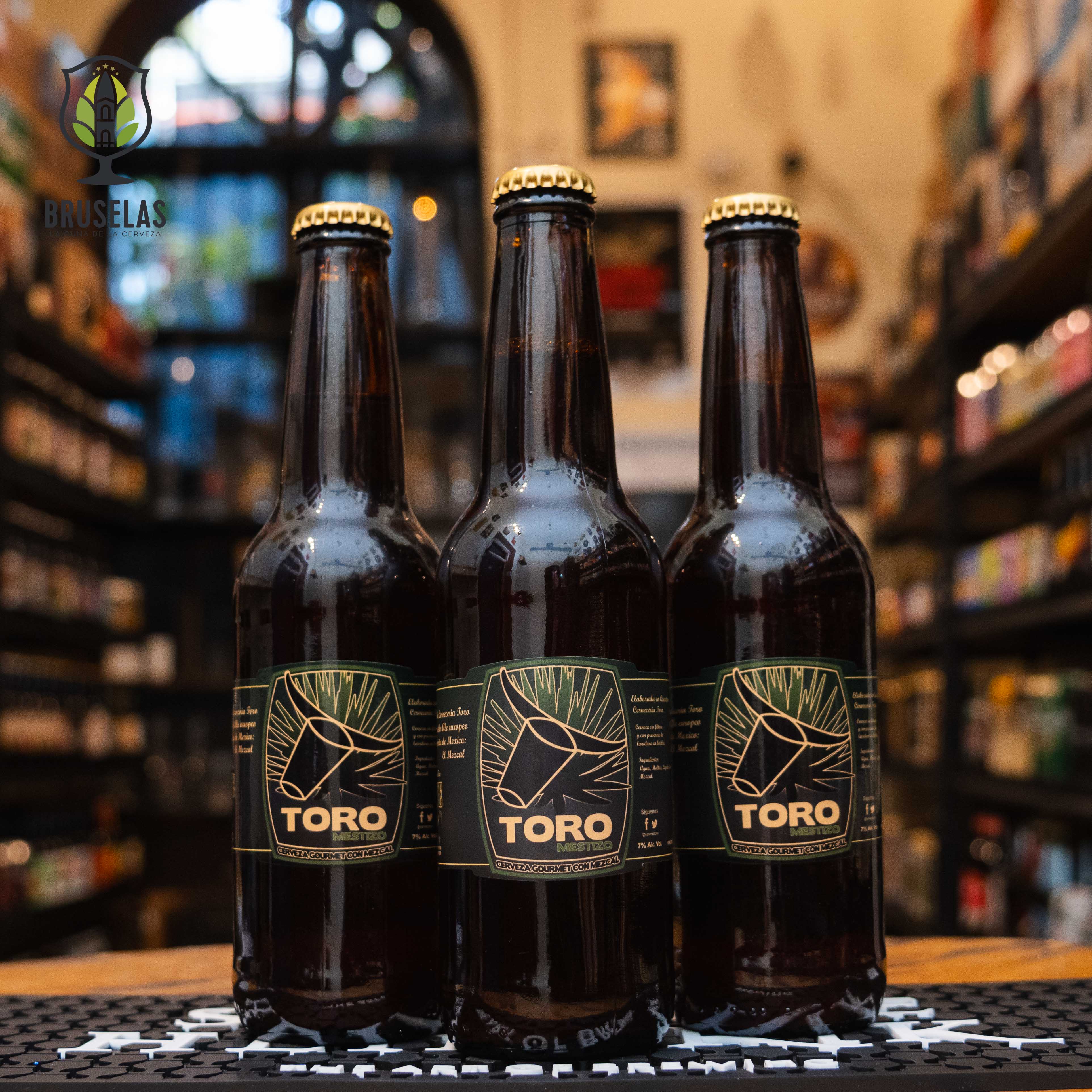 Botella de Toro Mestizo, una Agave Ale artesanal de Cervecería Toro, Querétaro, México. La etiqueta es negra con líneas doradas que forman un toro estilizado. ABV de 7.0%. Sabor ahumado con notas de mezcal, miel y coriandro. Ideal para acompañar carnes ahumadas, mole y botanas picantes. El diseño refleja una estética sofisticada y fuerte, evocando la cultura mexicana.