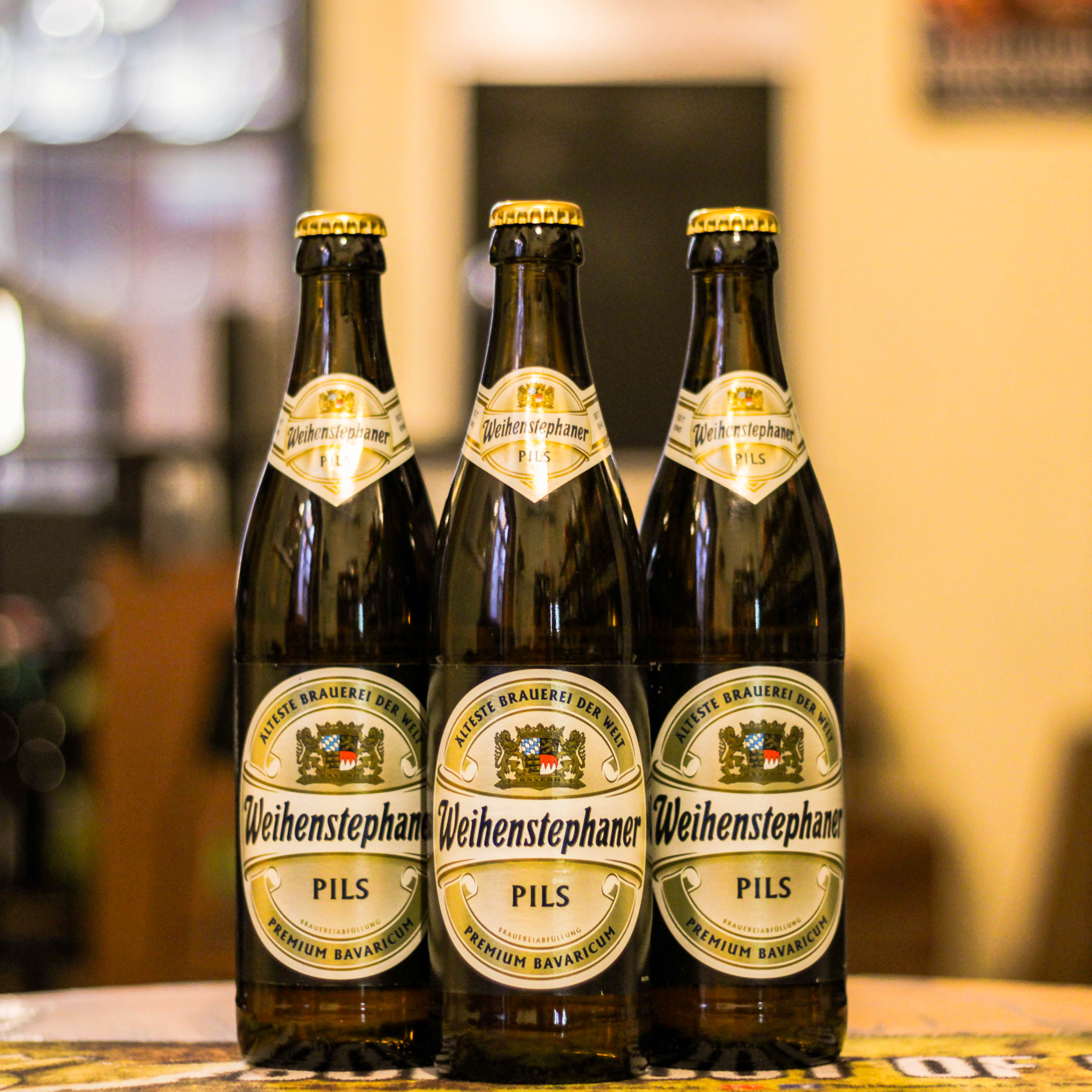 Botella de Weihenstephaner Pils, una pilsner bávara de la cervecería Weihenstephan en Alemania. La etiqueta presenta el escudo dorado con detalles en azul y blanco. ABV de 5.1% y 30 IBU, con un sabor equilibrado entre malta y lúpulo y aroma floral de lúpulos de Hallertau. Ideal con quesos suaves, pescados y carpaccio. Refleja la herencia de la cervecería más antigua del mundo.