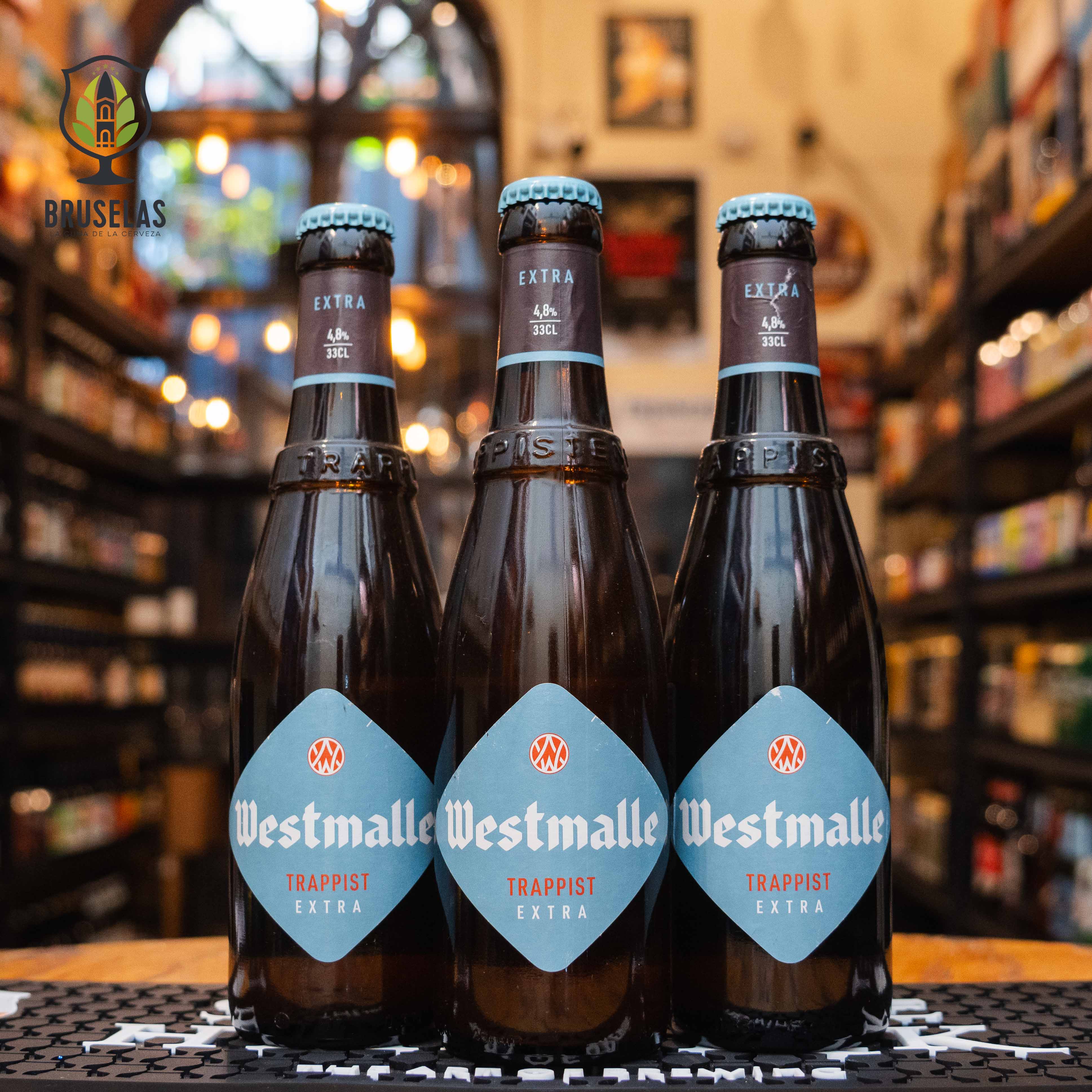 Botella de Westmalle Extra, una cerveza trapense belga de estilo Belgian Enkel/Patersbier. La etiqueta es azul claro con el logo trapense. ABV de 4.8%. Sabor fresco y afrutado con notas herbales y un amargor suave. Ideal para acompañar comidas ligeras, pescados y ensaladas. El diseño refleja la tradición y simplicidad de esta cerveza creada para el consumo de los monjes.