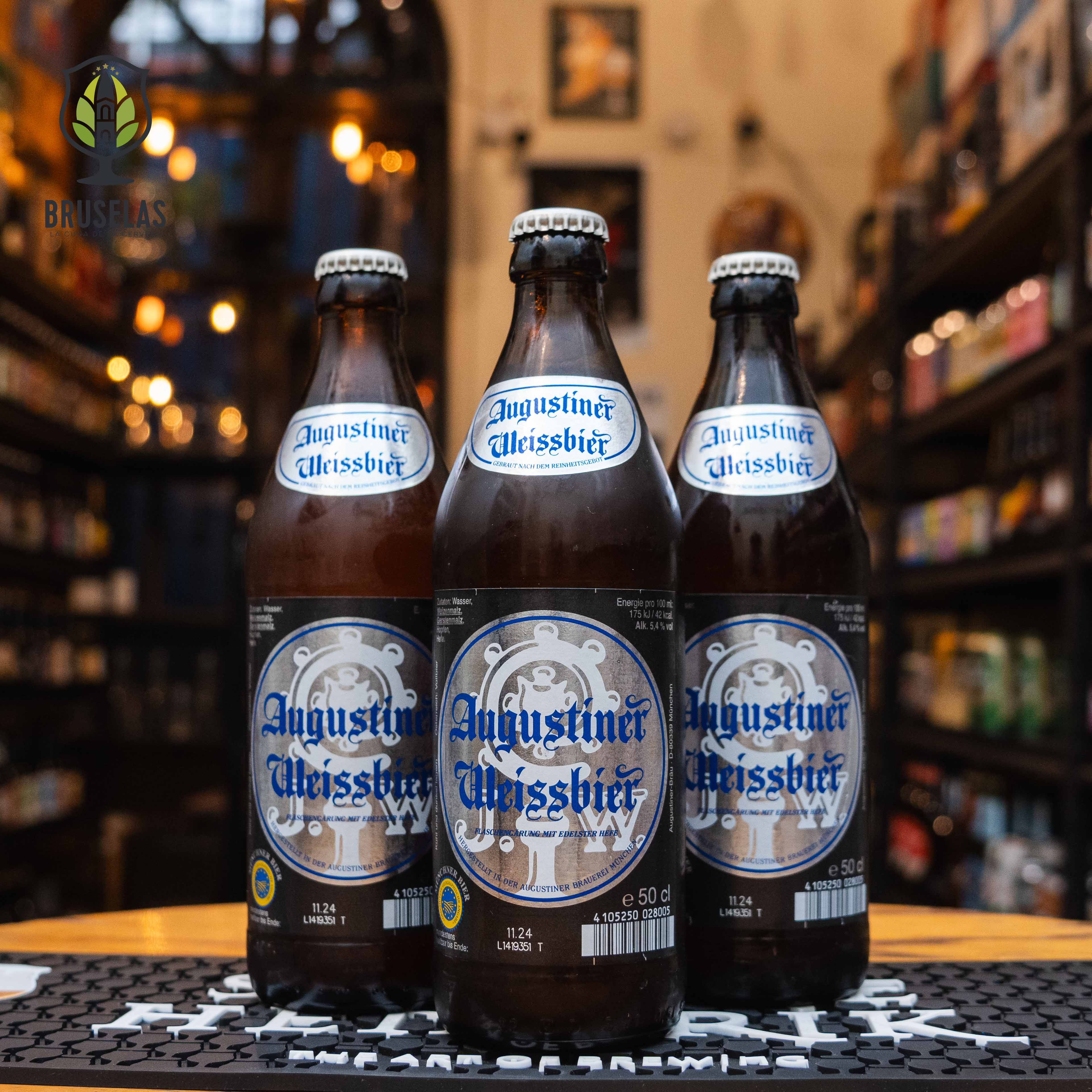 Botella de Augustiner Weissbier, una Hefeweizen de Augustiner-Bräu, elaborada en Múnich, Alemania. La etiqueta tiene un diseño en tonos plateados y azules, con un logotipo central que incluye un emblema de la cervecería. El nombre de la cerveza está en letras góticas azules. Presenta un ABV de 5.4% y un perfil de sabor con notas de plátano, clavo y ligeros toques cítricos. Ideal para acompañar ensaladas frescas, mariscos y pretzels. El diseño de la botella refleja un estilo clásico y tradicional bávaro. 