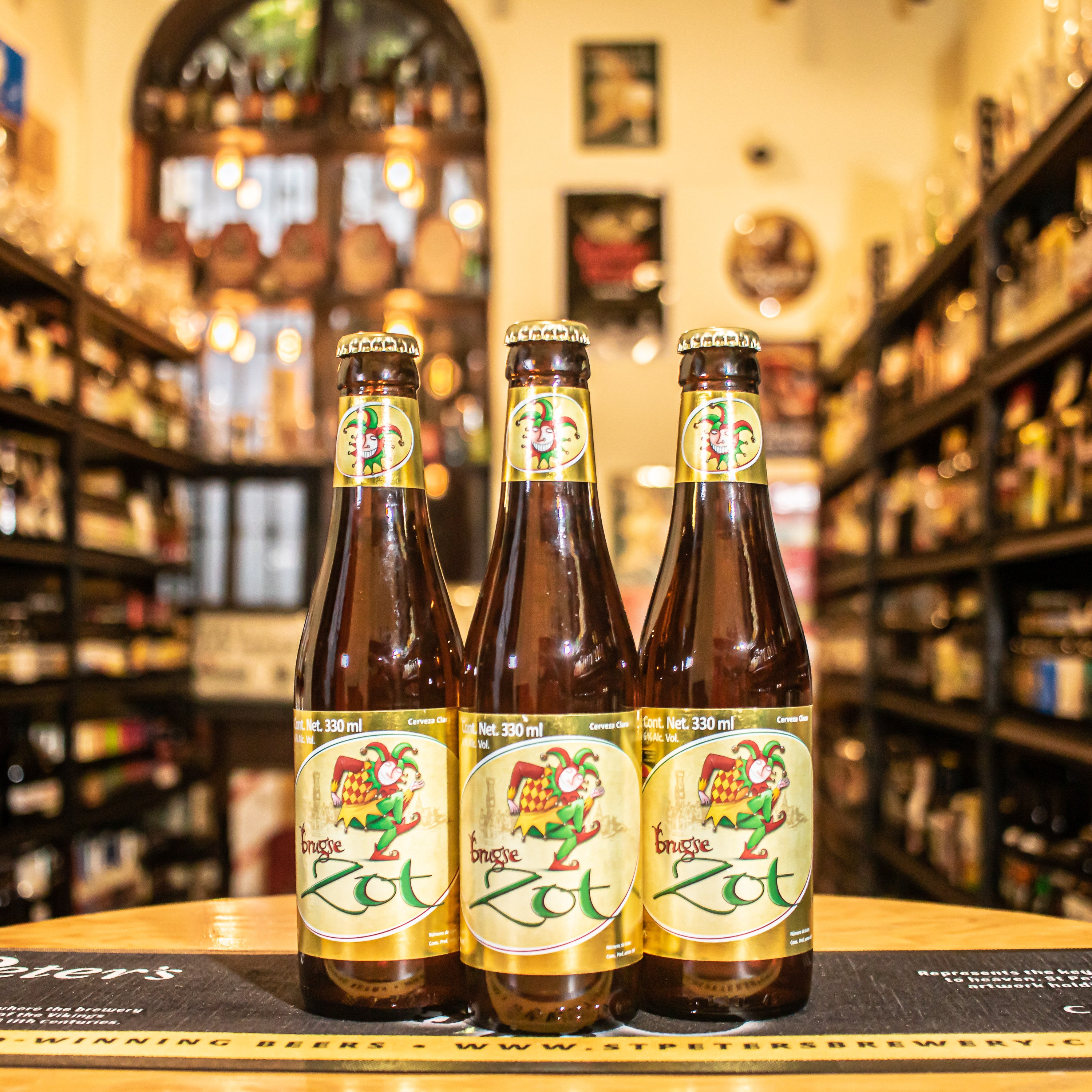 Botella de Brugse Zot Blond, una Belgian Blond Ale de la cervecería De Halve Maan, elaborada en Brujas, Bélgica. La etiqueta presenta un diseño tradicional con un bufón, destacando su origen histórico. Con un ABV de 6% y 23 IBU, ofrece aromas frutales y especiados con notas cítricas. Su sabor es equilibrado, con un amargor moderado y un final seco. Ideal para acompañar cocina mediterránea, pescados, mariscos y postres de frutos rojos. Su diseño refleja la rica tradición cervecera belga.