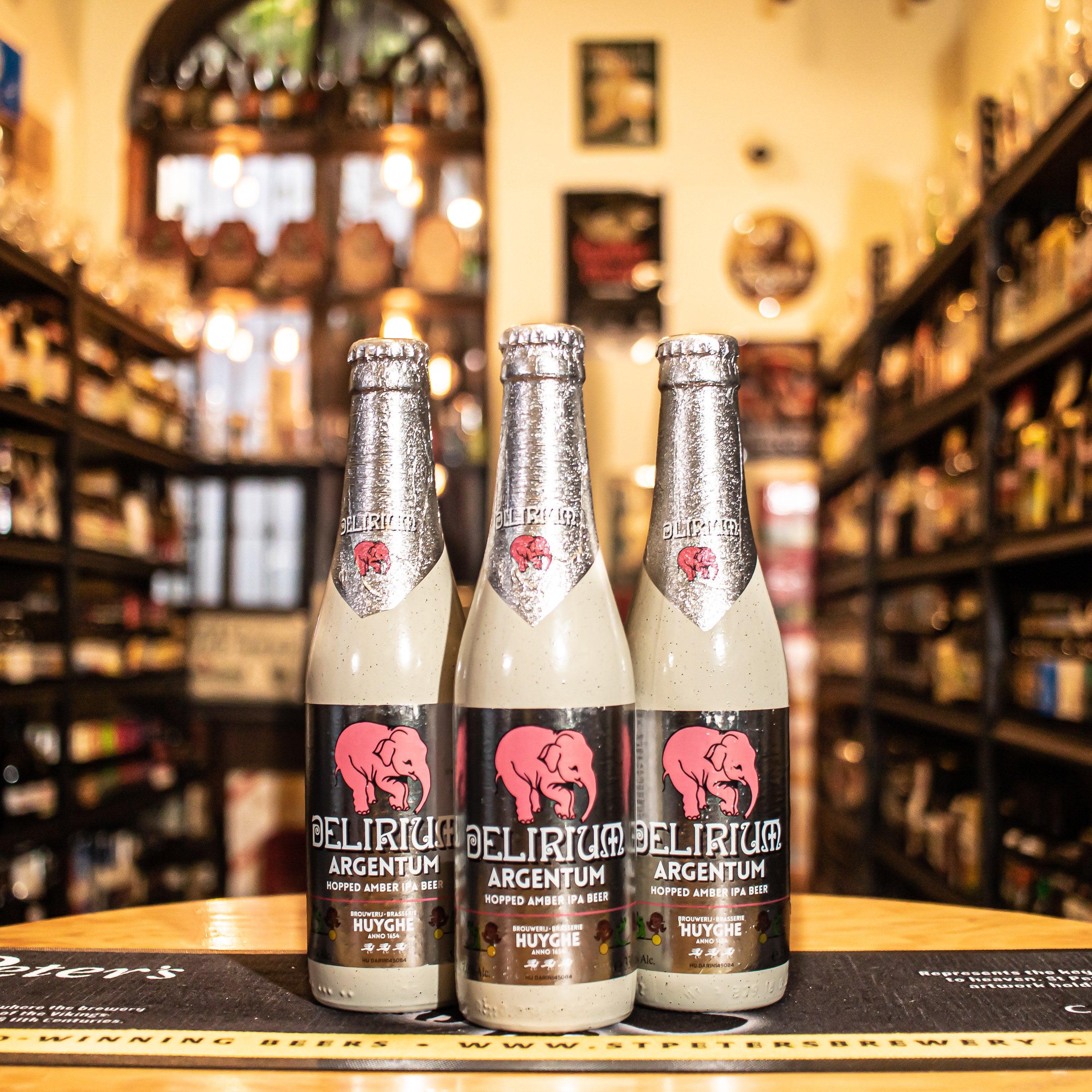 Botella de Delirium Argentum, una Belgian IPA de Brouwerij Huyghe, creada para celebrar el 25º aniversario de Delirium Tremens. Con un ABV de 7% y 49 IBU, presenta un color ámbar rojizo y espuma densa crema. Sus aromas incluyen especias, cítricos y caramelo. En boca, equilibra la dulzura del caramelo con amargor cítrico del lúpulo, con un final fresco y amargo. Ideal para carnes a la parrilla, platos especiados y quesos maduros.