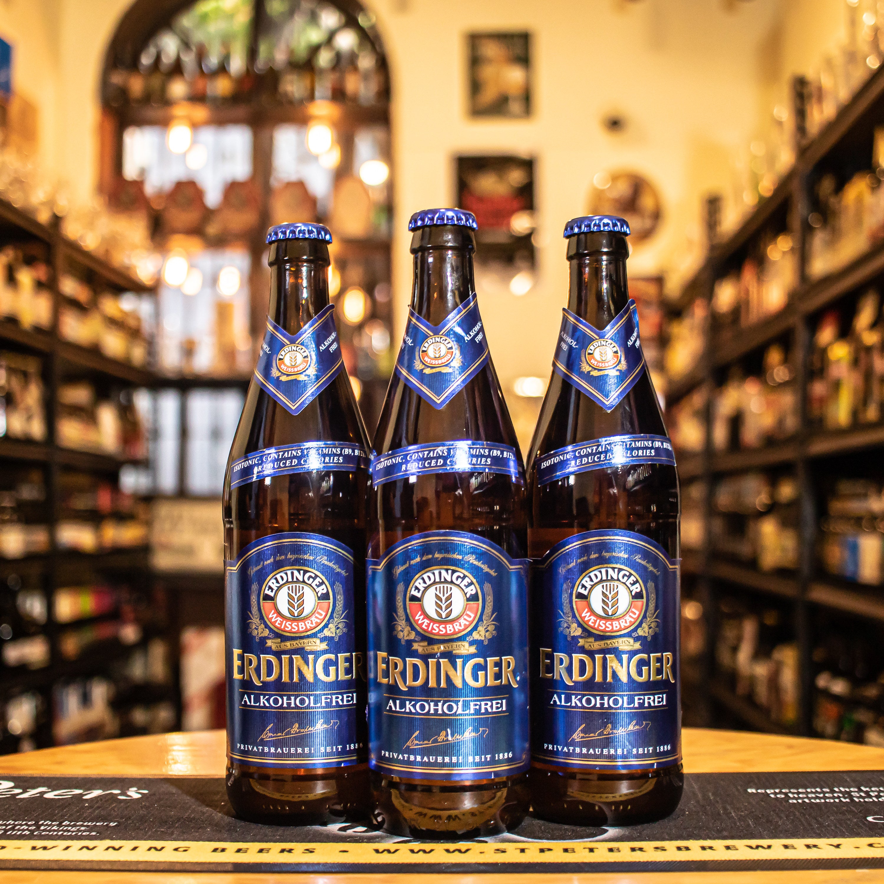 Botella de Erdinger Weissbier Alkoholfrei, una cerveza de trigo sin alcohol de Erdinger Weissbräu, Bavaria. Con menos de 0.5% ABV, destaca por su perfil isotónico, ideal para rehidratación tras el ejercicio. Su sabor pleno y refrescante emula a las cervezas con alcohol, con notas de malta especiada, caramelo y un toque frutal. Es ligera, con un suave amargor y acidez, perfecta para acompañar ensaladas, pescados o disfrutar sola.