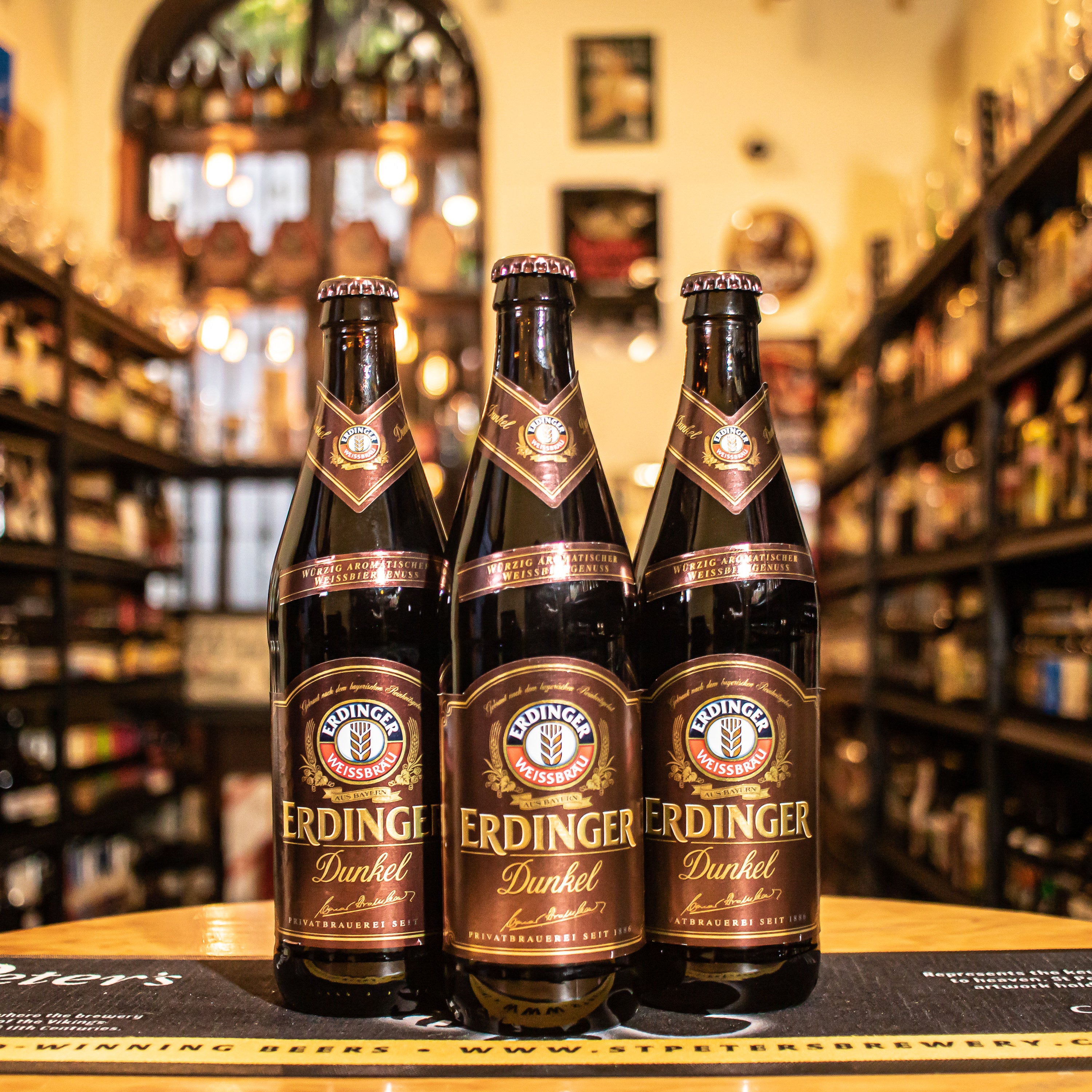 Botella de Erdinger Weissbier Dunkel, una cerveza de trigo oscura de la cervecería alemana Erdinger Weissbräu. Con un ABV de 5.3%, se caracteriza por su color marrón oscuro y espuma densa beige. Ofrece aromas a malta tostada, caramelo, café, clavo y plátano. En boca, predominan sabores de maltas tostadas, chocolate y especias, con un final seco y ligeramente amargo. Ideal para acompañar carnes a la parrilla, mariscos y platos intensos.