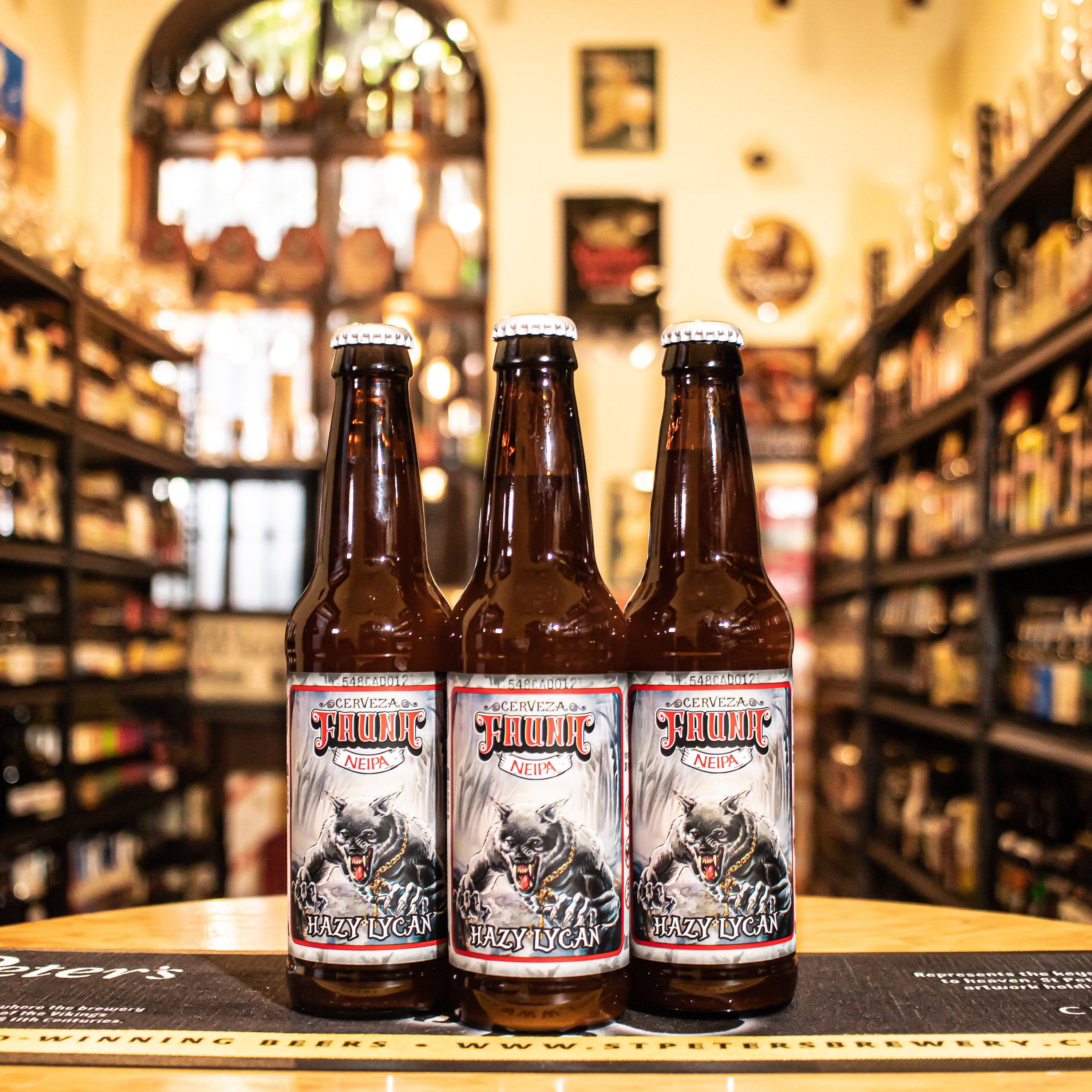 Botella de Hazy Lycan, una New England IPA de cervecería Fauna. La etiqueta muestra un lobo estilizado con tonos oscuros y detalles plateados. ABV de 7.0% e IBU de 38. Ofrece aromas a toronja, naranja y durazno, con sabores a frutas tropicales y un amargor moderado. Ideal para acompañar hamburguesas y tacos. Su diseño moderno refleja el carácter audaz y afrutado de esta cerveza.