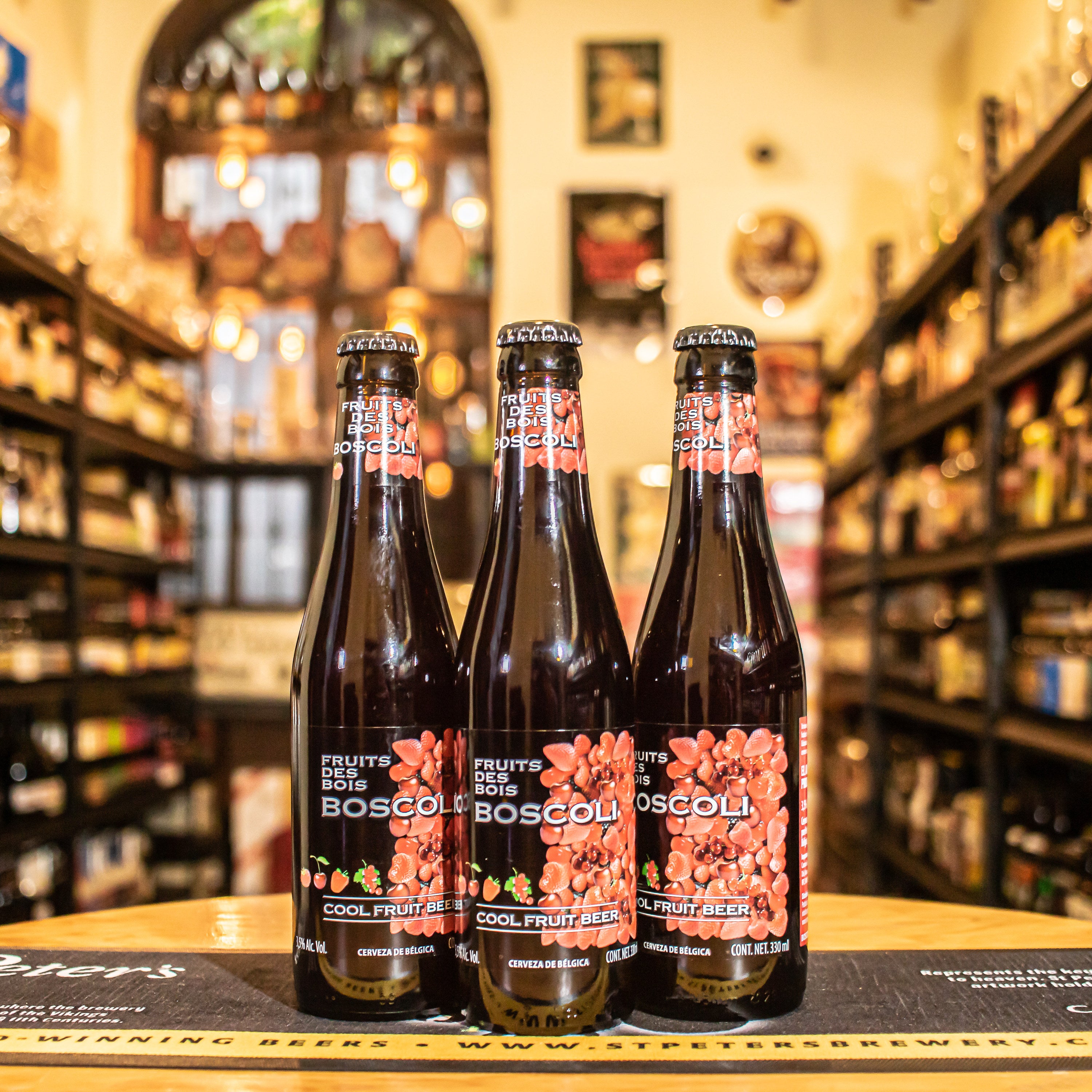 Botella de Boscoli Fruit de Bois, una Fruit Beer belga de Brouwerij Het Anker. La etiqueta presenta un diseño colorido que refleja su carácter afrutado. Con un ABV de 3.5%, esta cerveza ofrece aromas dulces de frambuesa, grosella y mora, con un sabor dulce y frutal equilibrado por una suave acidez. Ideal para acompañar postres, ensaladas de frutas y quesos suaves, o disfrutarla como una bebida refrescante. El diseño resalta su frescura y ligereza.