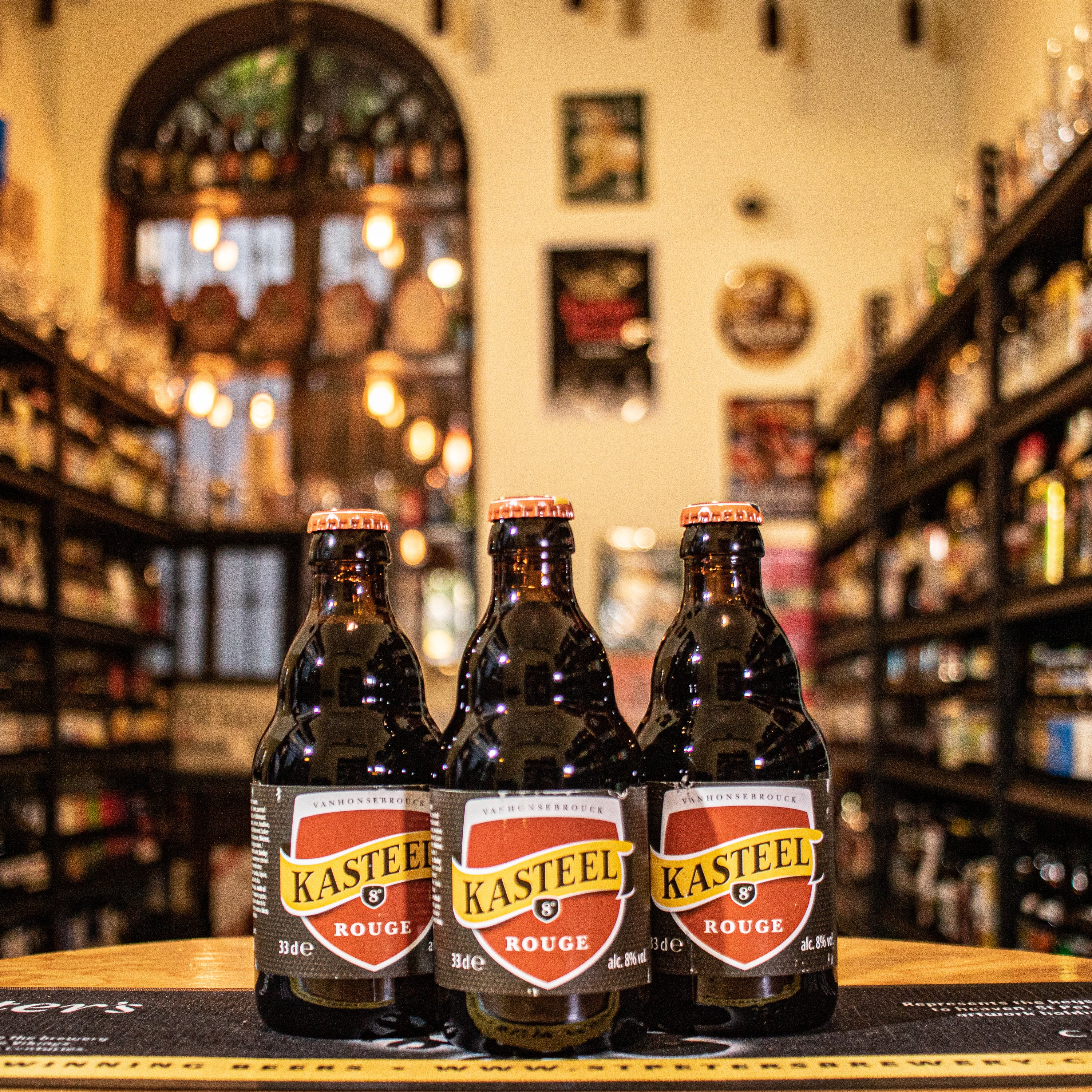 Botella de Kasteel Rouge, una Fruit Beer belga de Brouwerij Van Honsebrouck. Con un ABV de 8%, presenta un color rubí profundo. Elaborada con Belgian Strong Ale y jugo de cerezas frescas, ofrece aromas de cereza madura y un fondo dulce. Su sabor es dulce y afrutado, con un final ligeramente ácido. Ideal para maridar con postres de chocolate y tartas de frutas. El diseño refleja su carácter vibrante y afrutado.