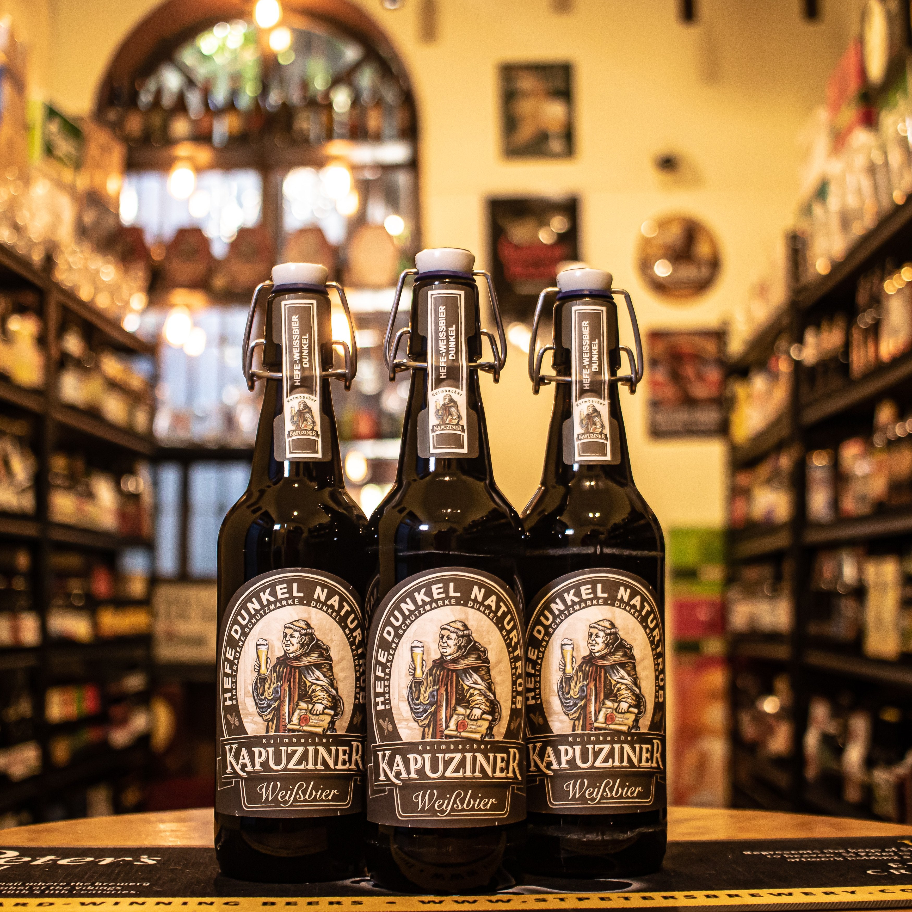 Botella de Kapuziner Schwarzbier, una cerveza oscura estilo Schwarzbier de la cervecería alemana Kapuziner. Con un ABV de 5.4%, presenta un color marrón profundo y espuma cremosa beige. Destacan sus aromas a malta tostada, caramelo y café. Su sabor es complejo, con un suave amargor y un final especiado. Ideal para maridar con carnes asadas, platos de caza o postres de chocolate como brownies.