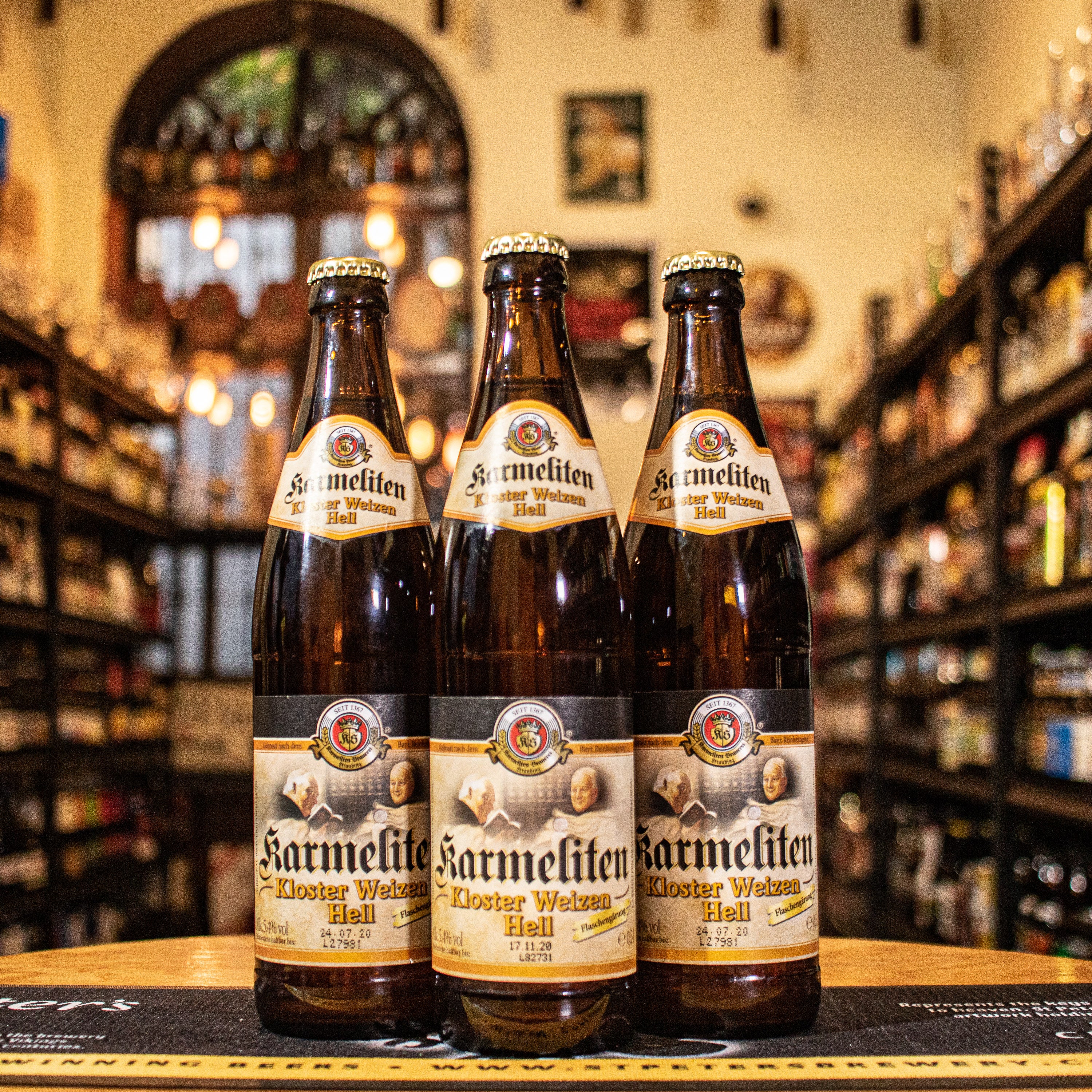 Botella de Karmeliten Kloster Weizen Hell, una cerveza de trigo estilo Weizen Hell de la cervecería alemana Karmeliten Brauerei. Con un ABV de 5.0%, tiene un color dorado pálido y una espuma cremosa. Ofrece aromas frutales de plátano y un toque de clavo. Su sabor es suave y balanceado, ideal para maridar con ensaladas, pescados y platos ligeros. El diseño refleja la frescura de esta clásica cerveza alemana.