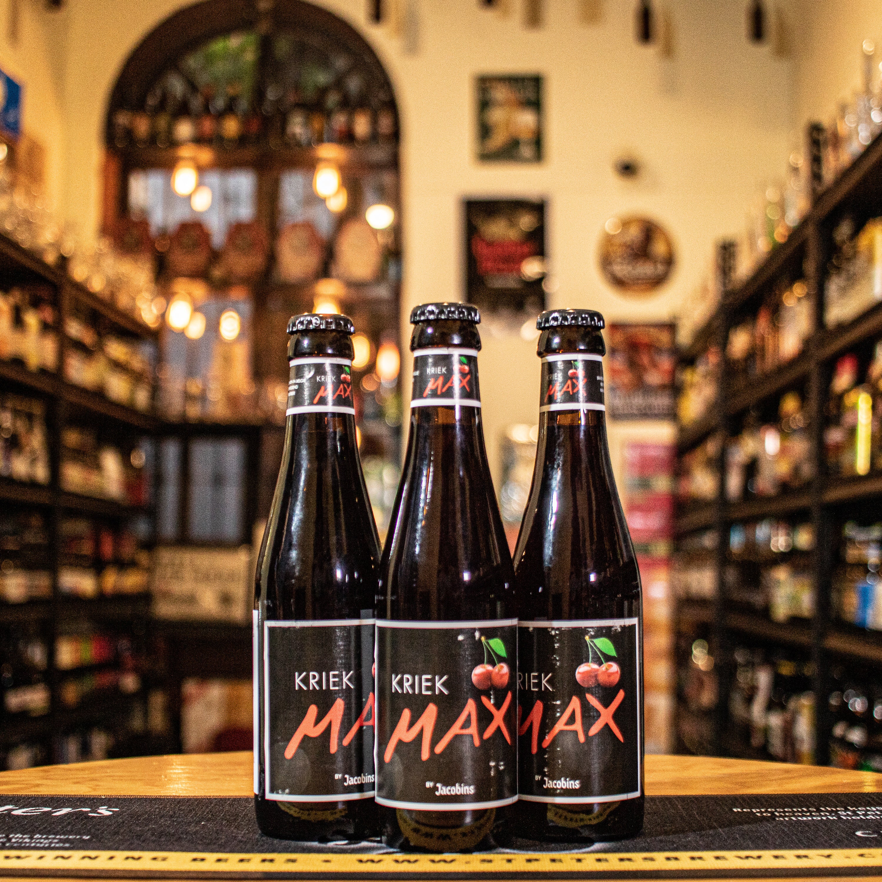 Botella de Kriek Max, una Fruit Beer belga de la cervecería Omer Vander Ghinste. Con un ABV de 3.2%, presenta un color rojo brillante y espuma ligera. Elaborada con cerezas frescas, ofrece aromas frutales y dulces. Su sabor es dulce y afrutado, con predominancia de cereza. Ideal para maridar con postres de frutas, quesos suaves y ensaladas frescas. El diseño refleja su carácter refrescante y vibrante.