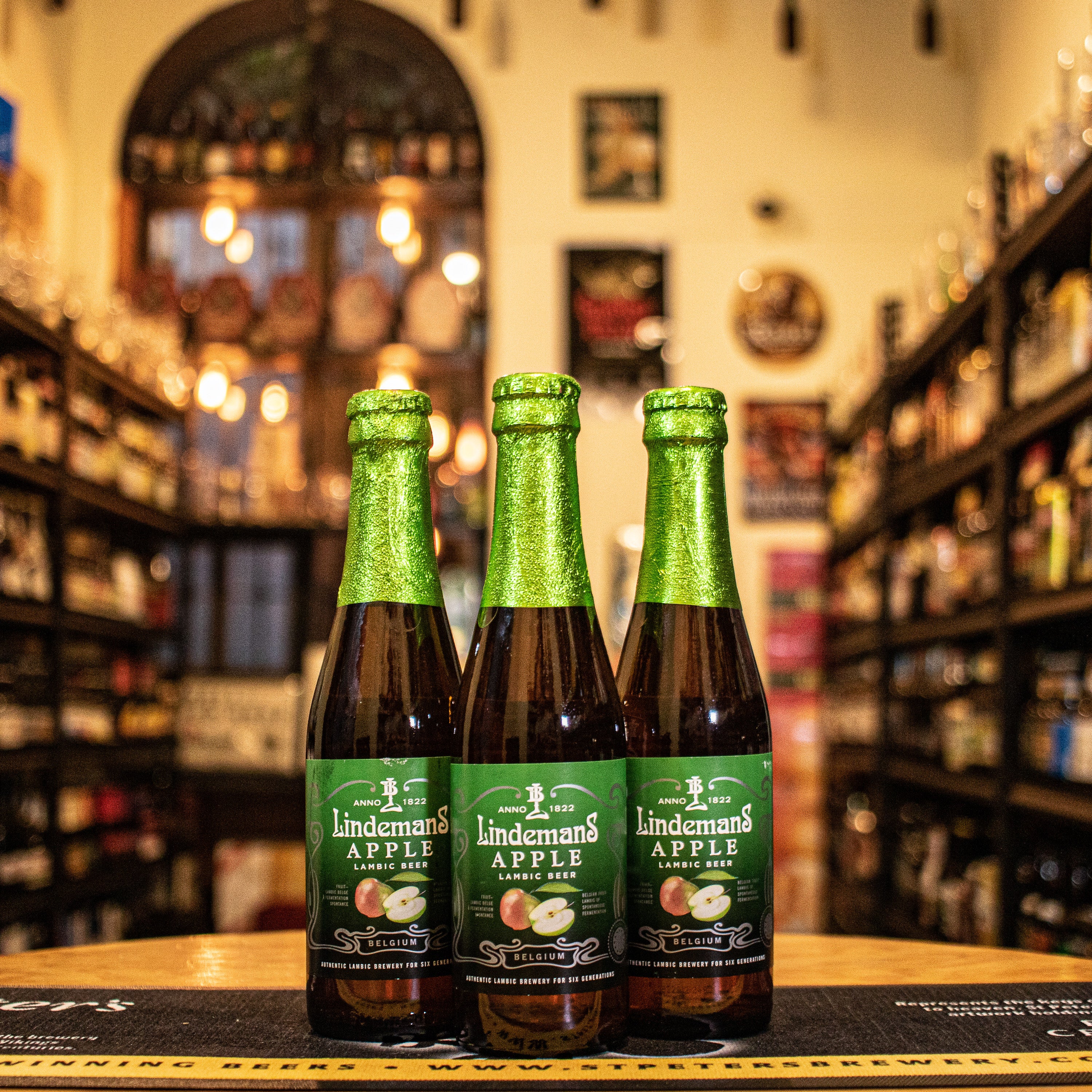 Botella de Lindemans Apple, una Fruit Lambic belga de Brouwerij Lindemans. Con un ABV de 3.5%, está elaborada con jugo de manzana verde. Ofrece aromas frescos y afrutados, con notas de manzana y un toque de levadura. Su sabor equilibra la acidez del lambic con la dulzura de la manzana, creando un final limpio y refrescante. Ideal para maridar con ensaladas ligeras, quesos suaves y postres de frutas.