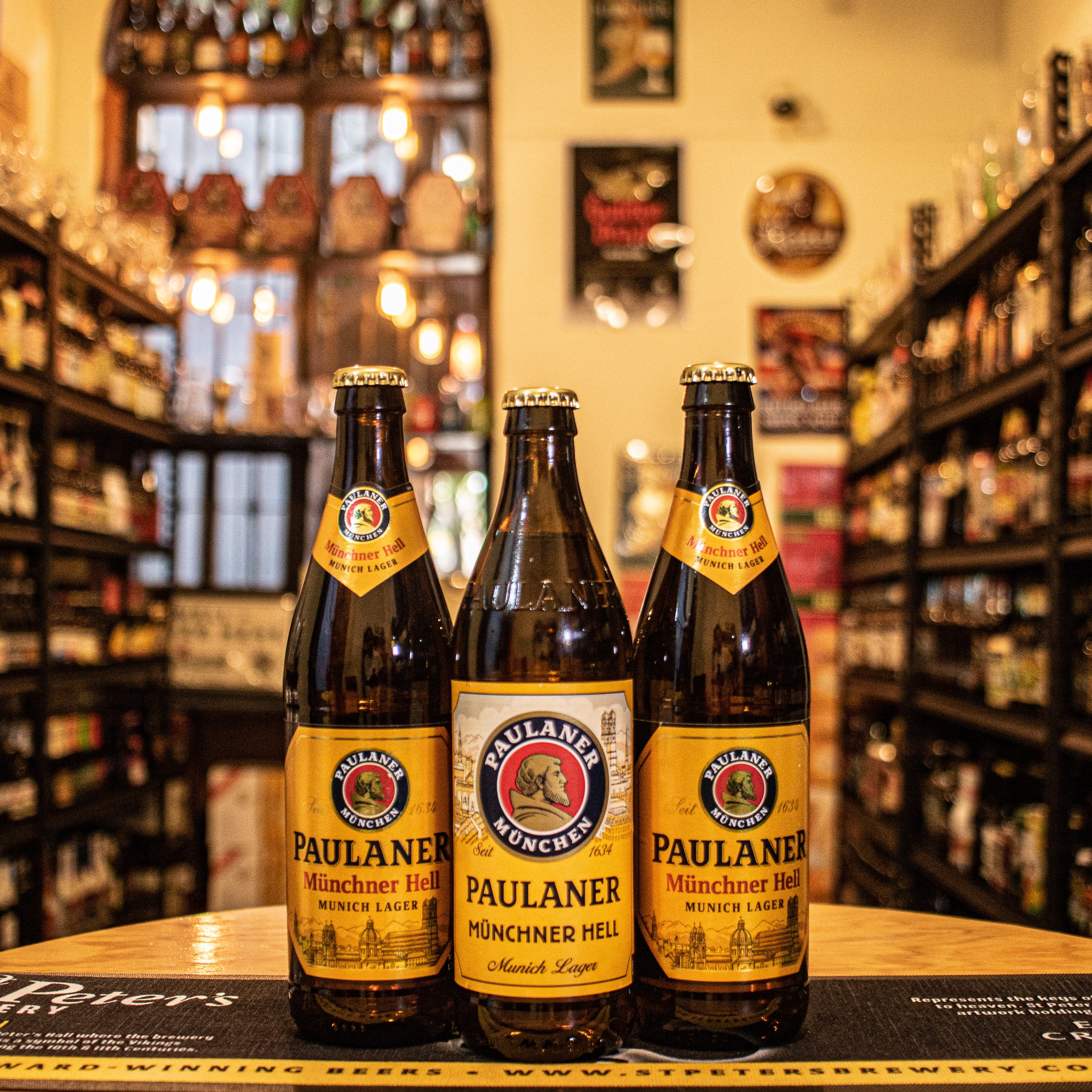 Botella de Paulaner Münchner Hell, una lager clara estilo Helles con un ABV de 4.9%, originaria de Múnich, Alemania. Presenta un color dorado brillante y espuma blanca persistente, con aromas de malta dulce y un toque de lúpulo. Su sabor es suave, equilibrando dulzor y amargor, con un final refrescante. Ideal para maridar con platos bávaros como salchichas y pretzels. Refleja la tradición cervecera bávara.